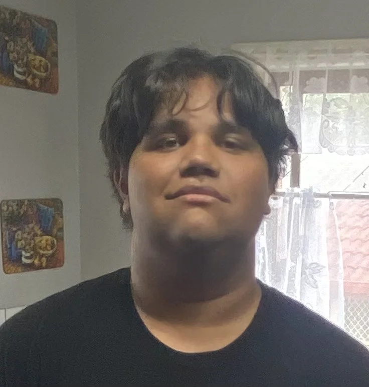 A young man with messy dark hair and a confident expression standing indoors near a window with lace curtains, with kitchen wall decorations in the background.