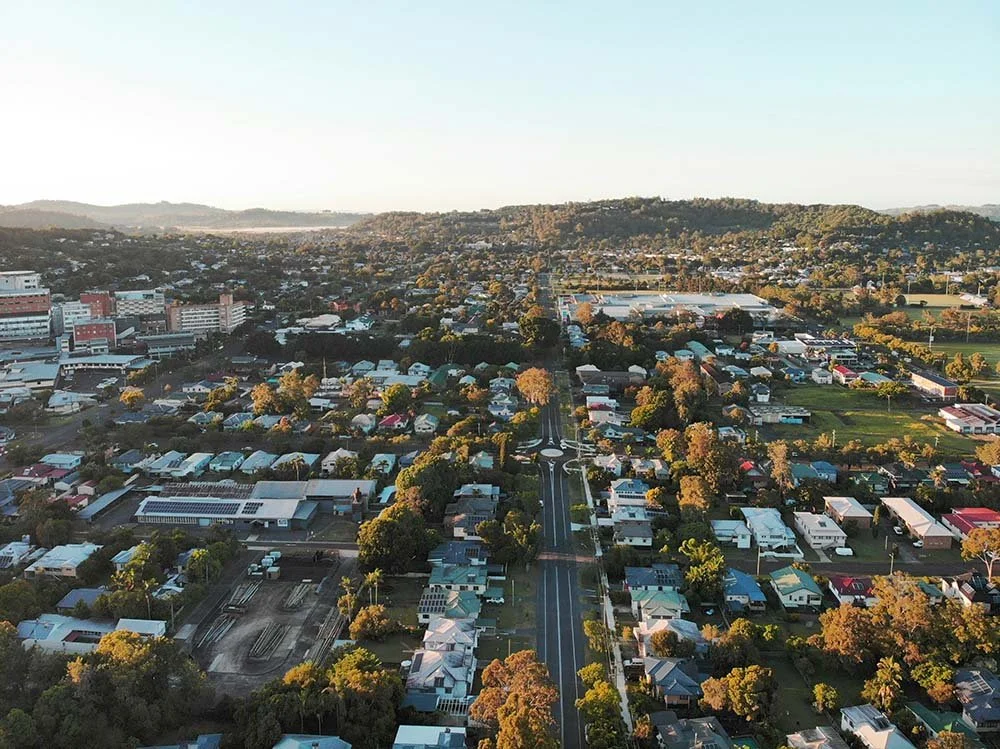 Aerial view of a suburban neighborhood with houses, trees, roads, and a distant cityscape.