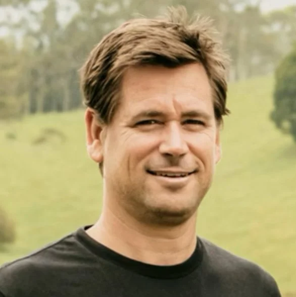 A man with brown hair smiling outdoors in a grassy area with trees in the background.