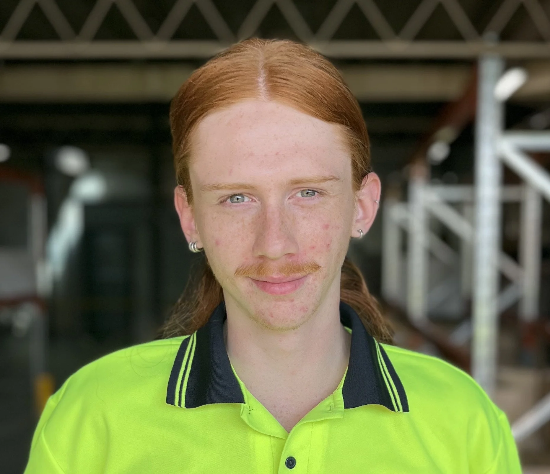 A young man with long red hair, light blue eyes, and a mustache, wearing a bright yellow safety shirt with black collar and accents, standing in front of an industrial or construction site background.