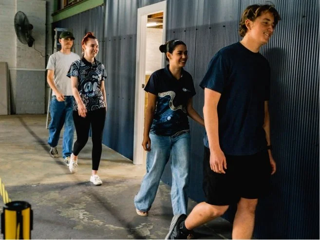 Four young people walking in a line inside a warehouse or industrial building, smiling and casually dressed.