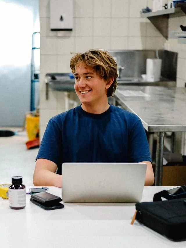 Smiling young man with wavy light brown hair wearing a blue T-shirt sitting at a table with a silver laptop, a small black wallet, a pair of glasses, a bottle of supplements, and a black bag in front of him in a kitchen or break room.