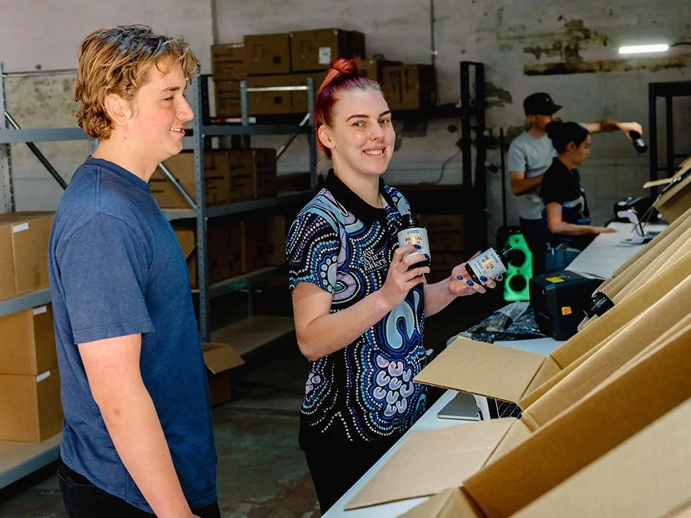 Two women in a warehouse, one smiling and holding canned goods, the other smiling and looking at her, with shelves of boxes and two other people working at a desk in the background.