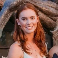 A young woman with red hair smiling outdoors in front of a large tree trunk.