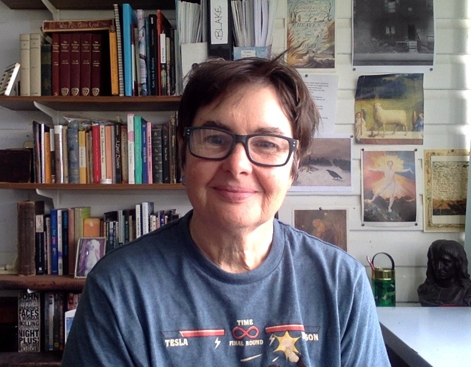 A woman with short dark hair and glasses smiling indoors, with bookshelves and decorative art on the wall behind her.