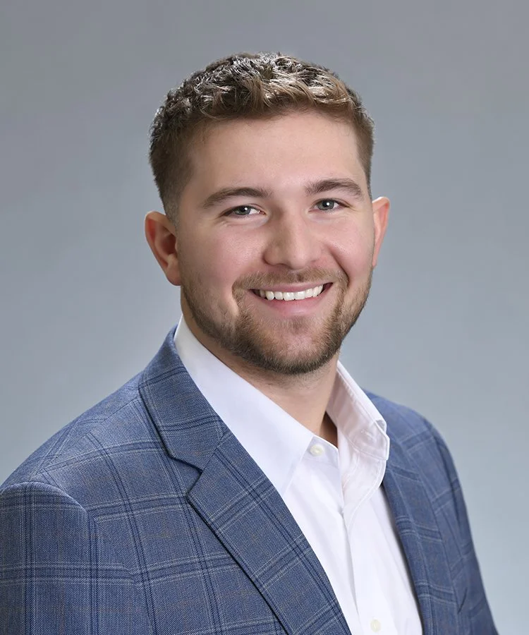 A young man with short, styled brown hair, a trimmed beard, wearing a blue plaid suit jacket and a white dress shirt, smiling against a neutral gray background.