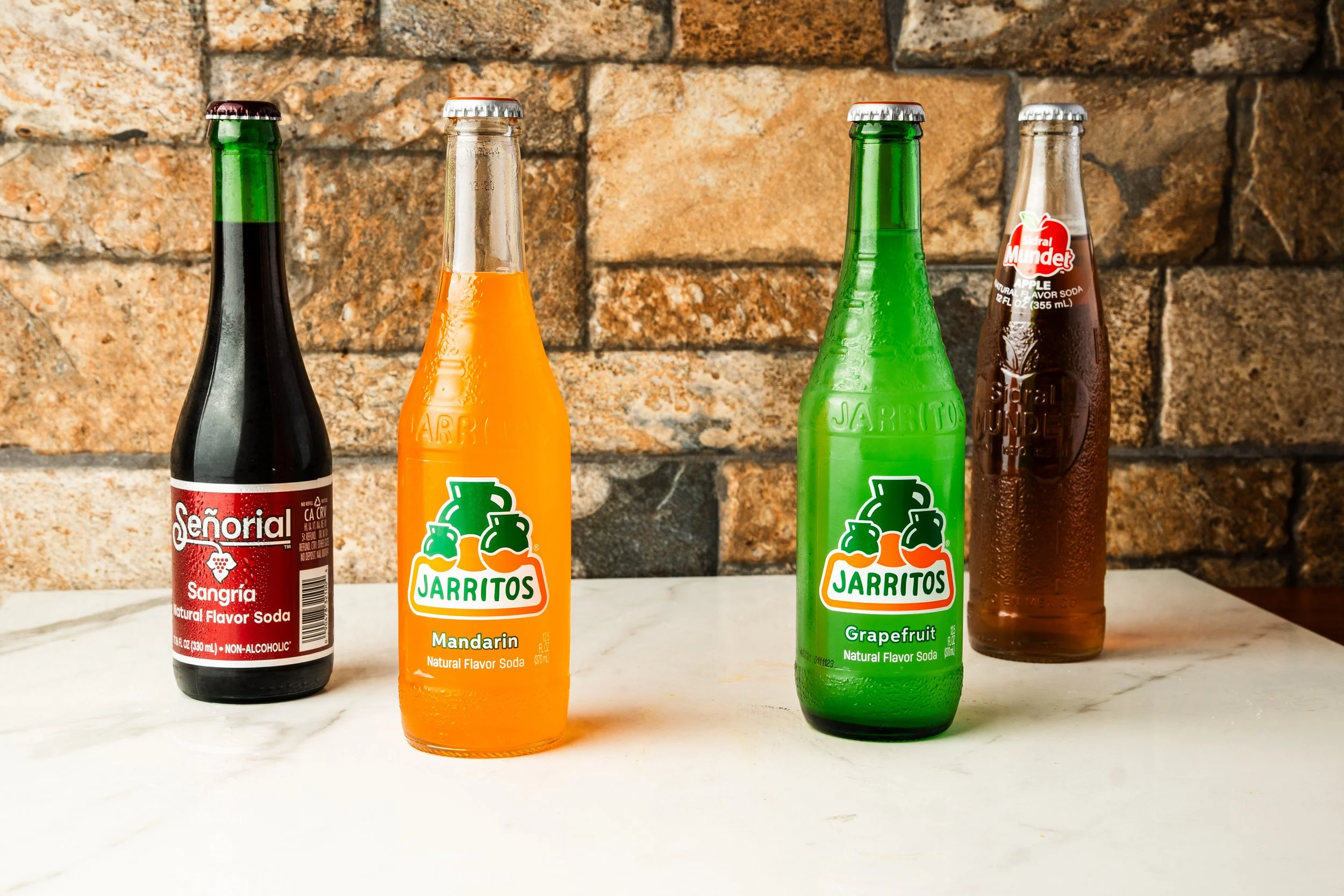 Four glass soda bottles on a white surface with a brick wall background. The bottles are Señoria Sangria, Jarritos Mandarin, Jarritos Grapefruit, and Mexican Coke.