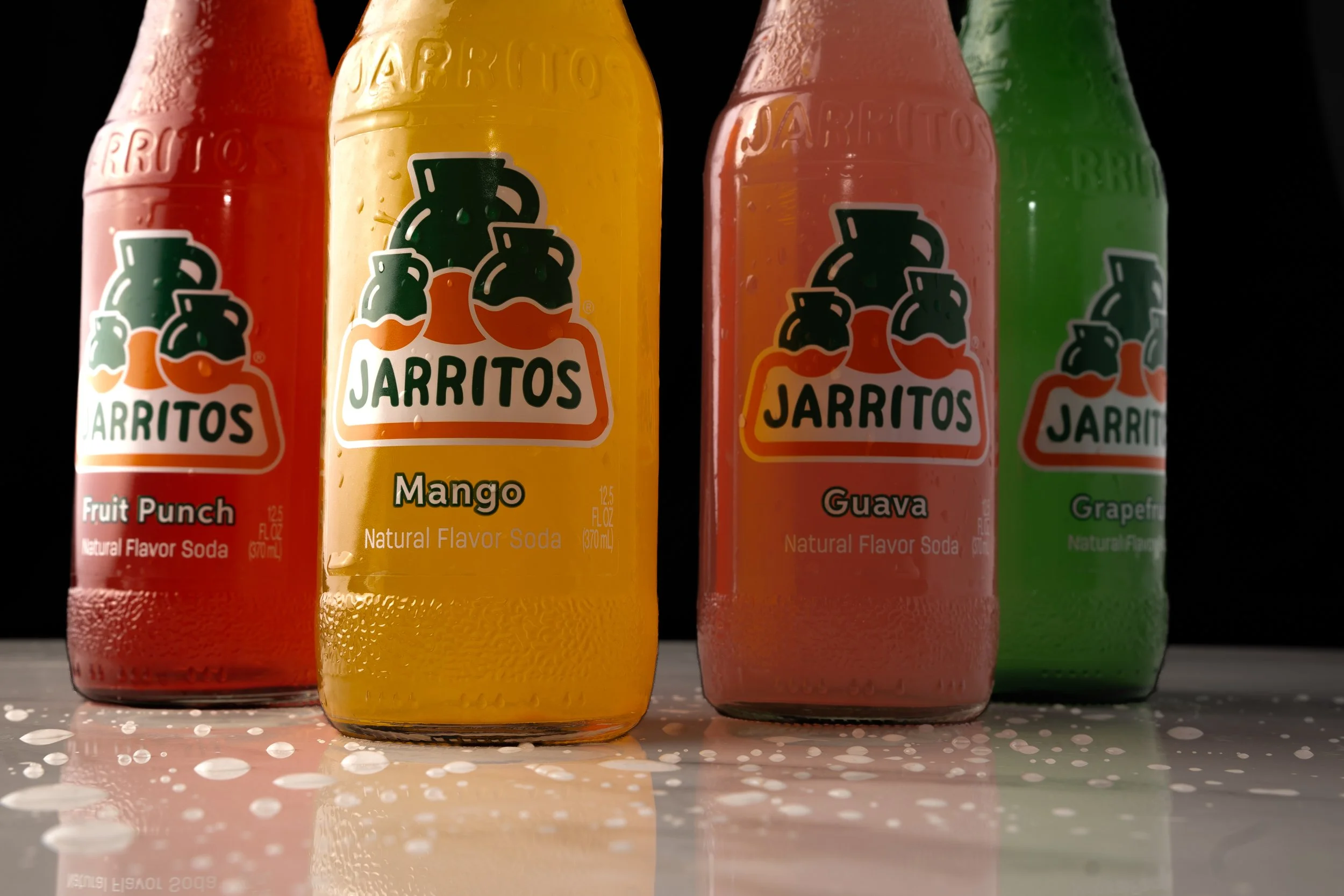 Four bottles of Jarritos flavored soda arranged in a row with condensation on the bottles. From left to right: Fruit Punch, Mango, Guava, and Grapefruit.