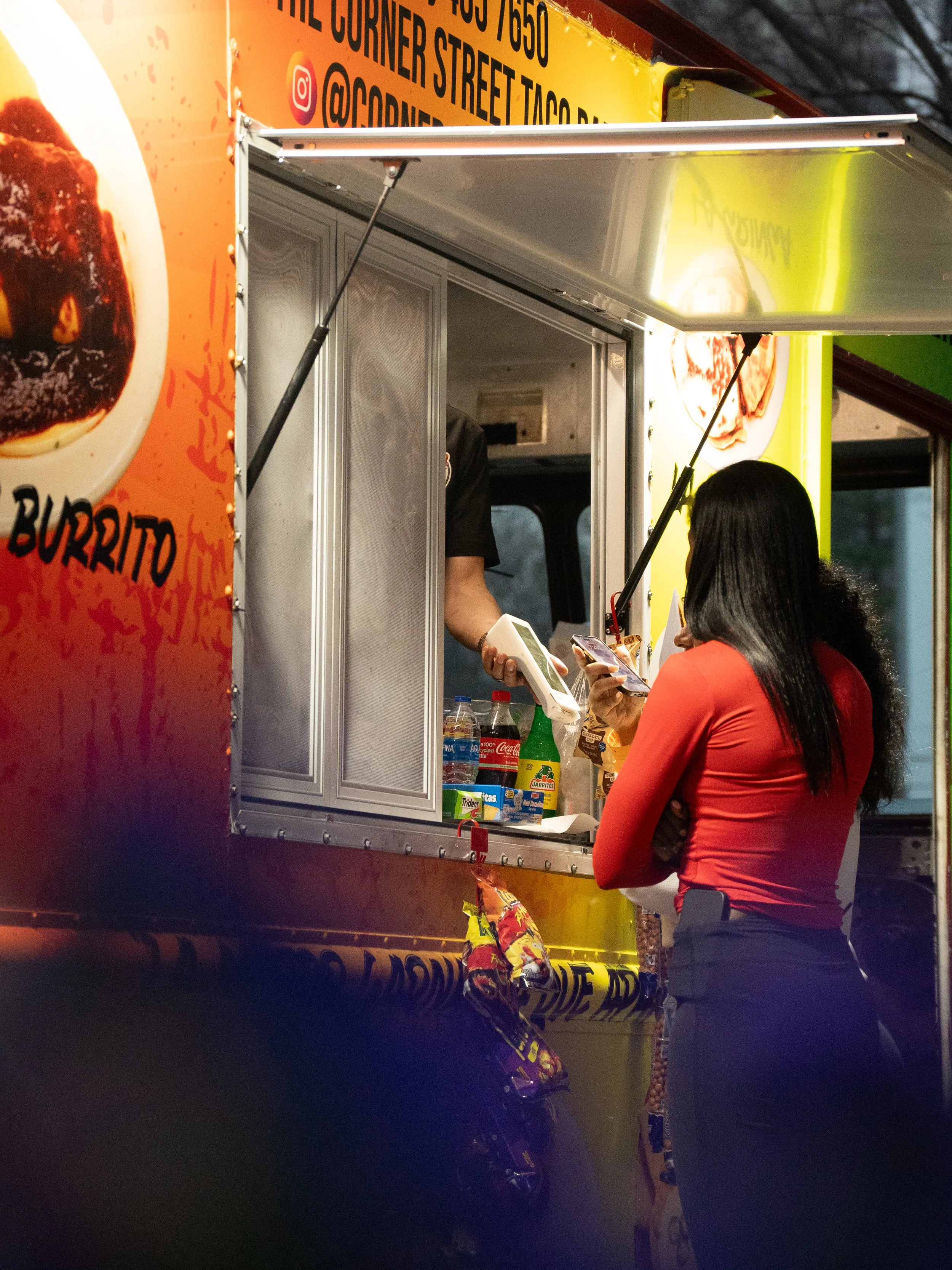 A woman with long black hair, wearing a red top and dark pants, is standing at a food truck counter, paying or ordering with a card. The food truck has orange and yellow colors with pictures of food, and a man is inside taking her order.