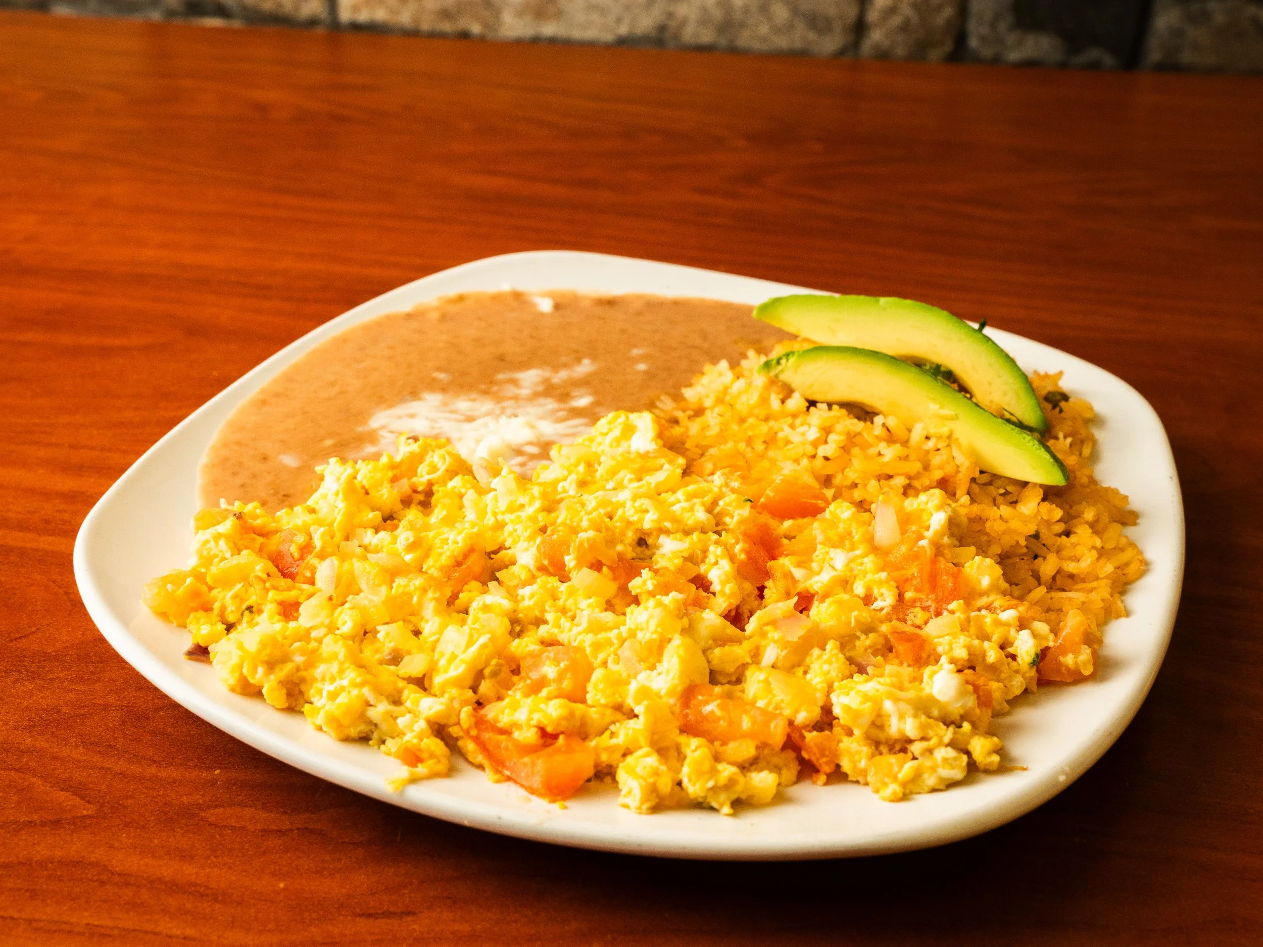 Plate of scrambled eggs with tomatoes, avocado slices, refried beans, and rice, on a white dish on a wooden table.