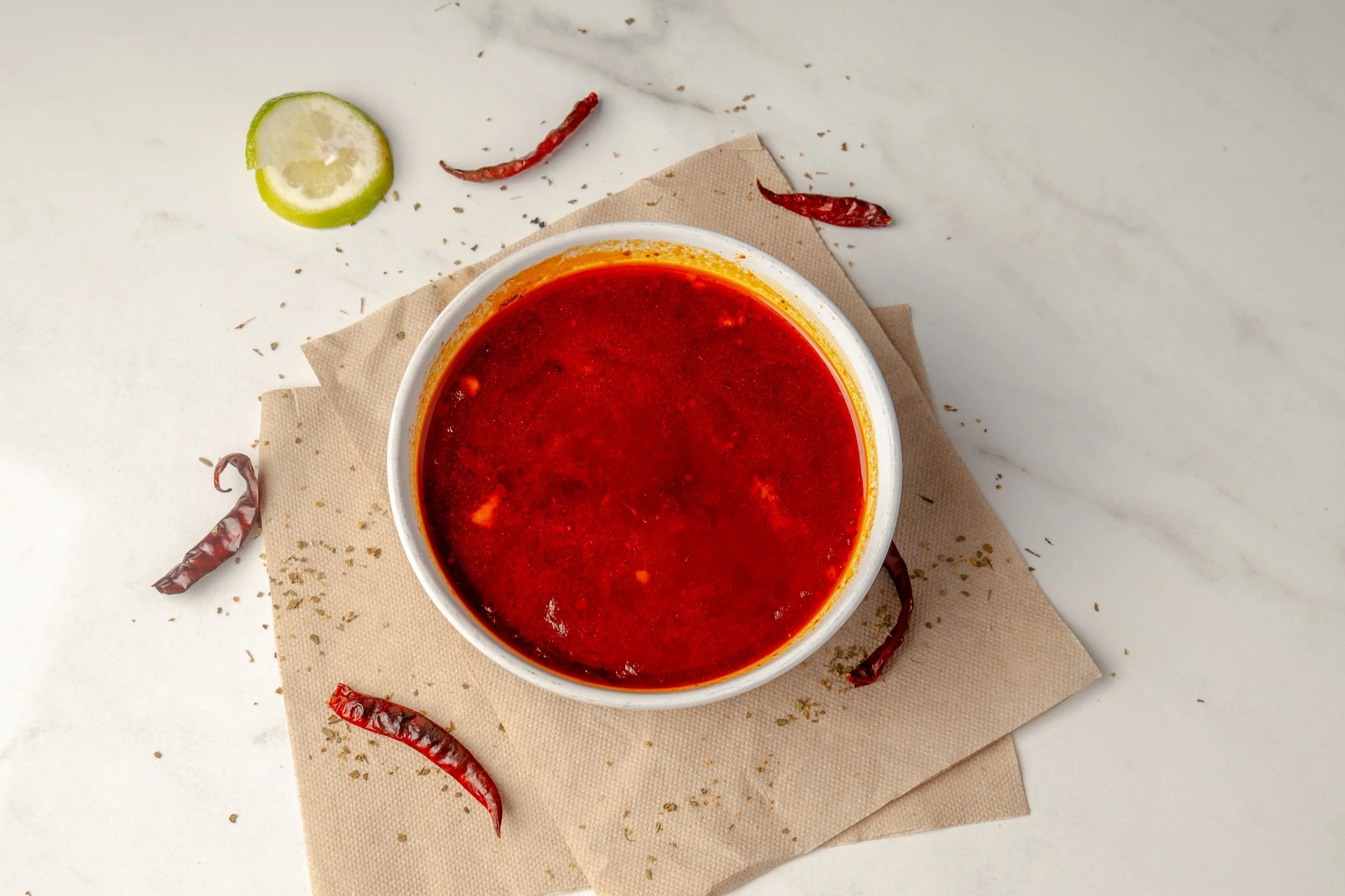 A bowl of Menudo soup with dried red chilies, a lime wedge, and scattered spices on a white marble surface.