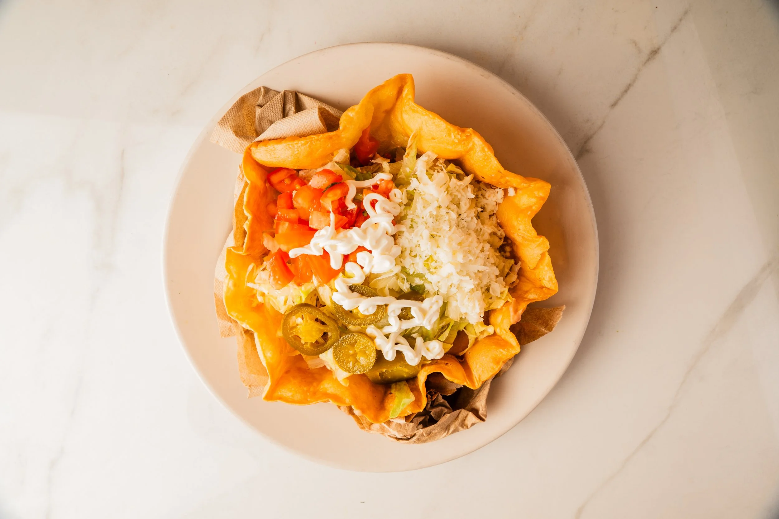 Taco shell filled with chopped tomatoes, shredded cheese, sliced jalapeños, lettuce, and topped with mayonnaise, on a white plate.