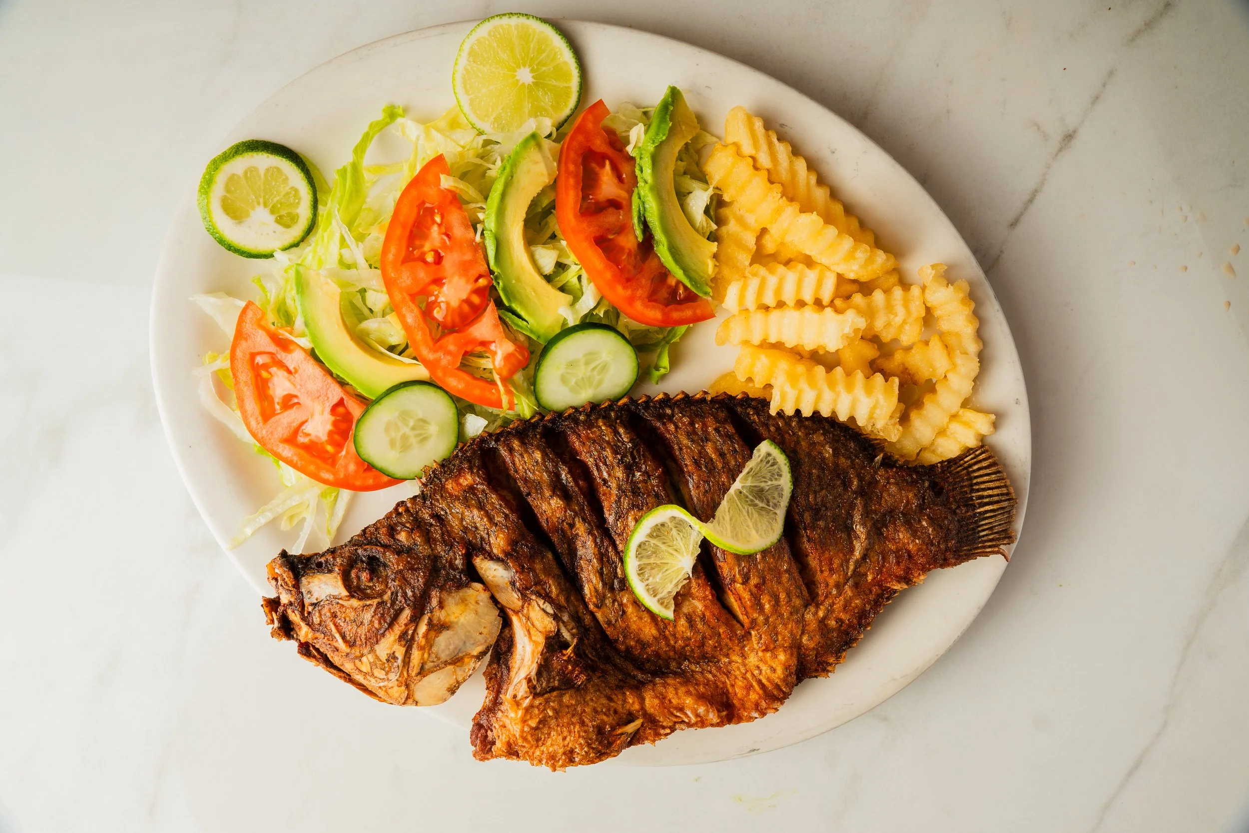 Fried whole fish with lemon slices, served with a side salad of tomatoes, cucumbers, avocado, lettuce, and lime slices, and a portion of crinkle-cut French fries on a white plate.