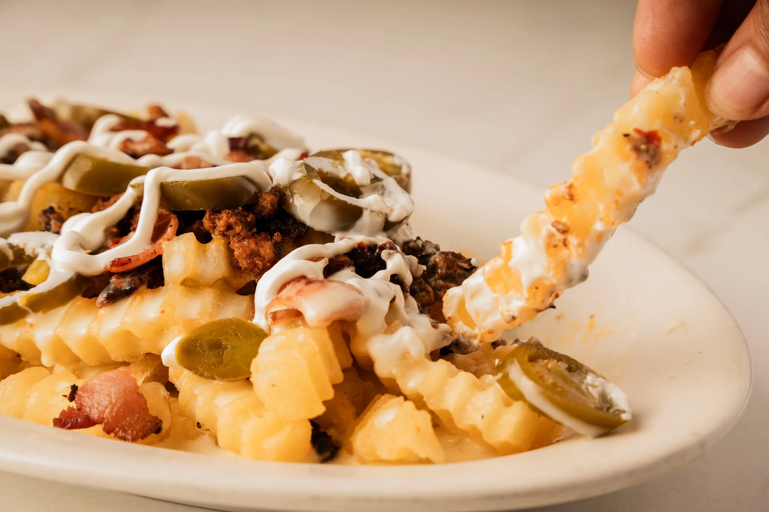 Close-up of a plate of loaded French fries topped with shredded cheese, jalapenos, bacon bits, ground beef, pickles, and drizzled with white sauce. A person's hand is holding a fry being dipped into a creamy cheese sauce.