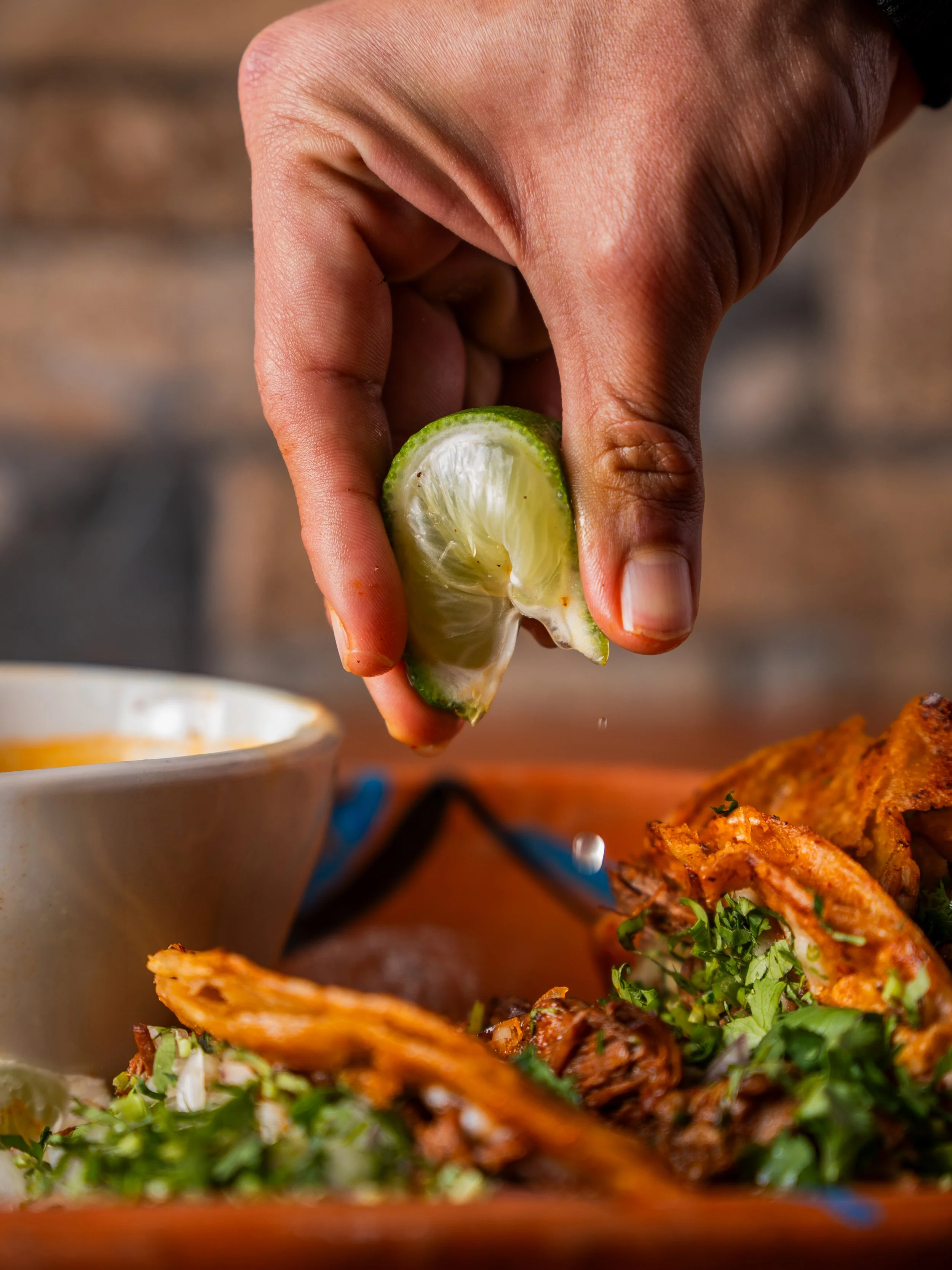 A hand squeezing a wedge of lime over a plate of Birria Tacos, with a bowl of Consome in the background.