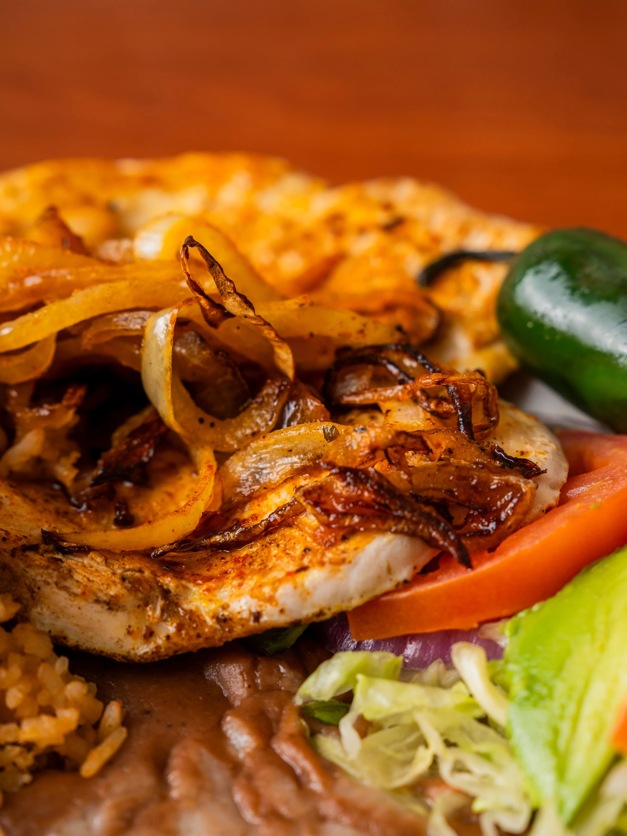 Close-up of grilled chicken breast topped with sautéed onions, served with sliced tomatoes, lettuce, and a jalapeño pepper.