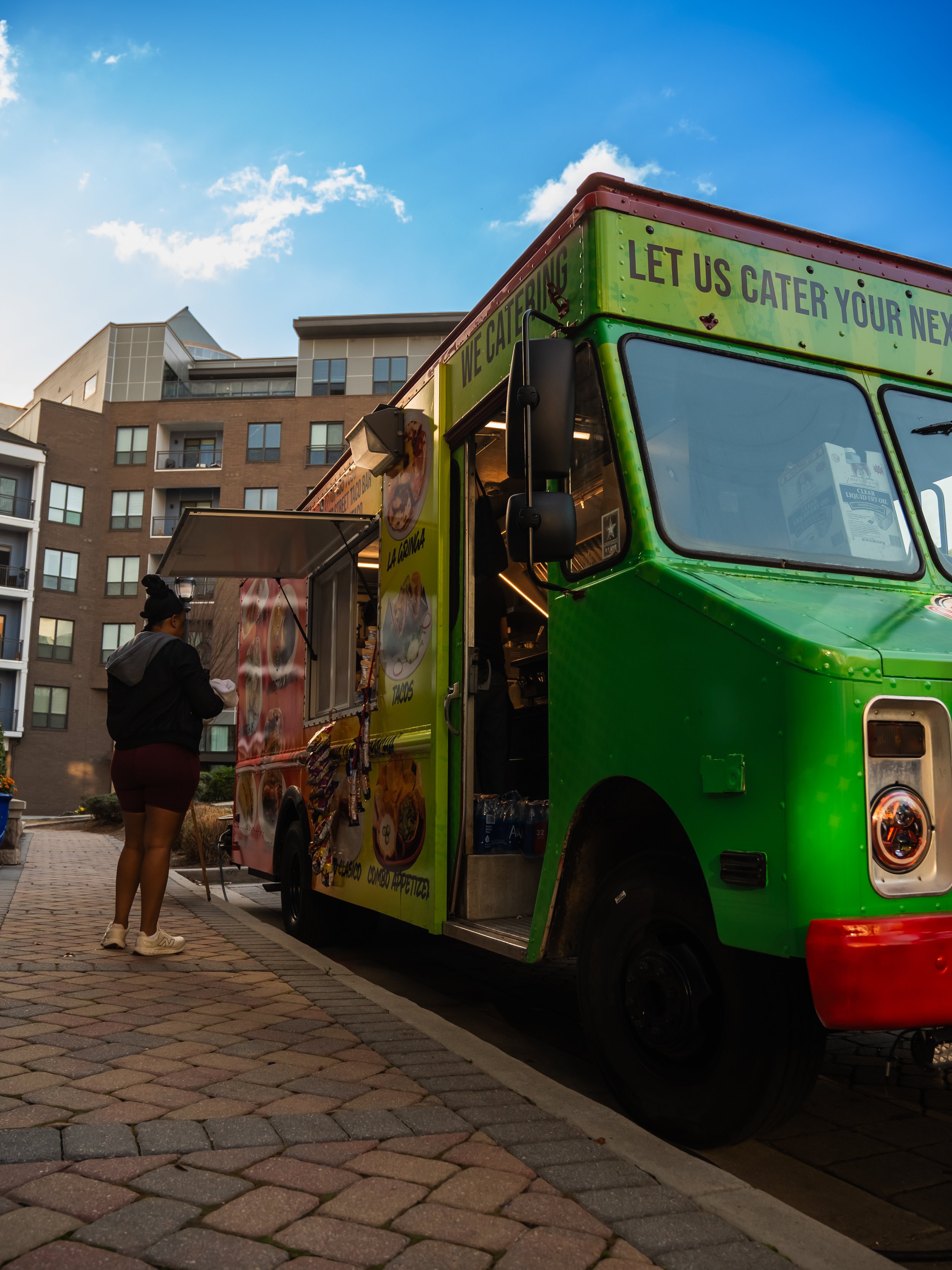 A woman placing an order at a green food truck with yellow accents, serving Mexican food, on a paved street with apartment buildings in the background during daytime.