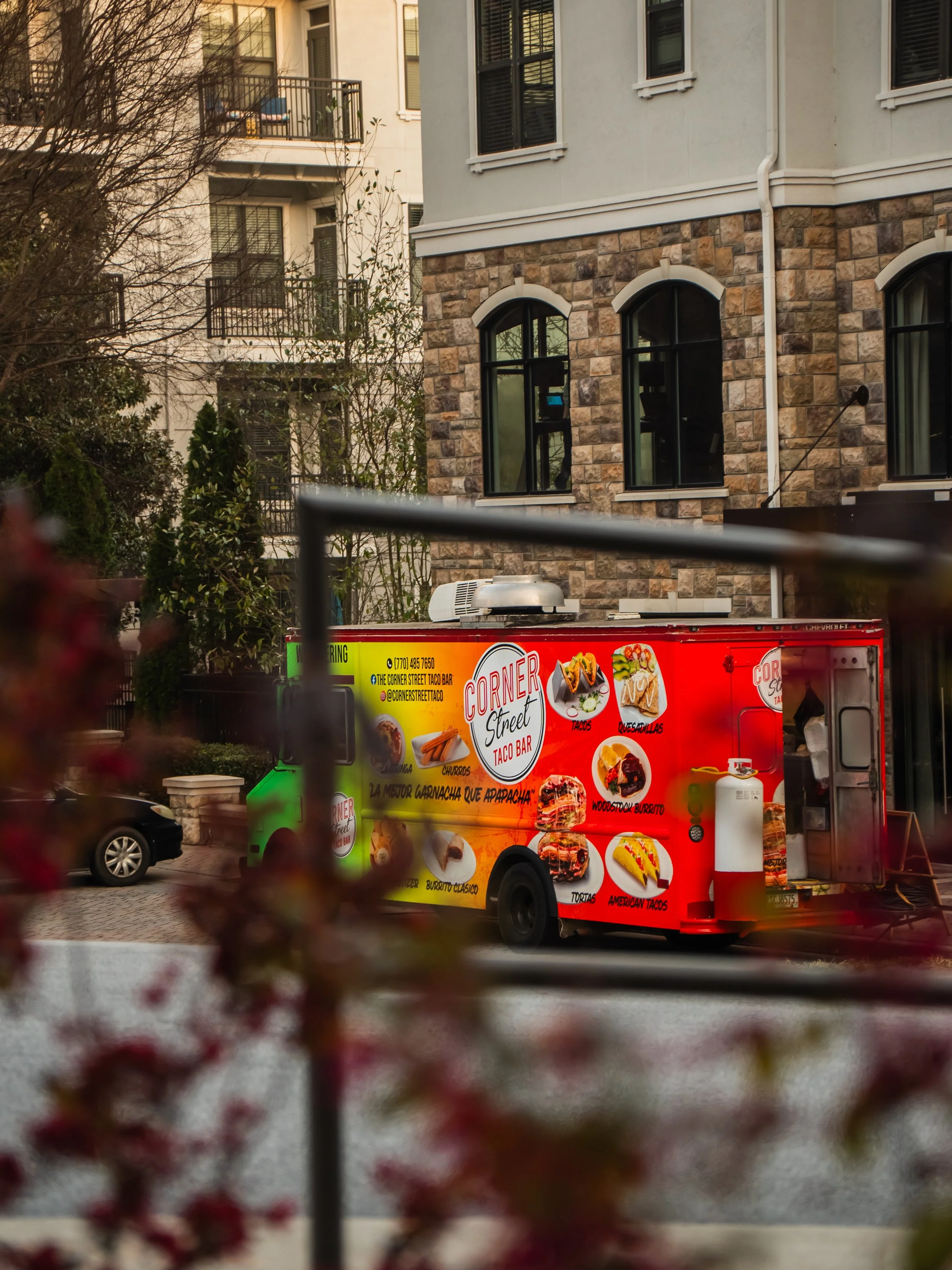 Red food truck parked on the street in front of a residential building with stone and white exterior walls. The truck has images of tacos, quesadillas, burritos, and American tacos, and the text 'Corner Street Taco Bar' and contact information.