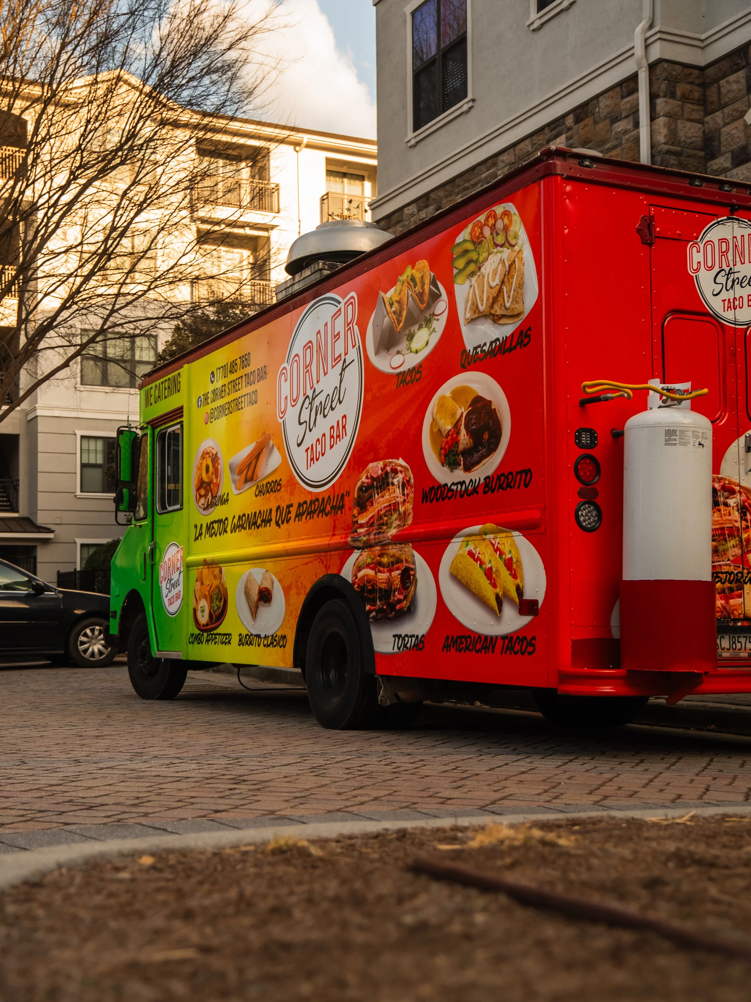 A red and green food truck named Corner Street Taco Bar, parked on a brick street in a residential area, featuring images of tacos, quesadillas, a burrito, and other Mexican dishes.