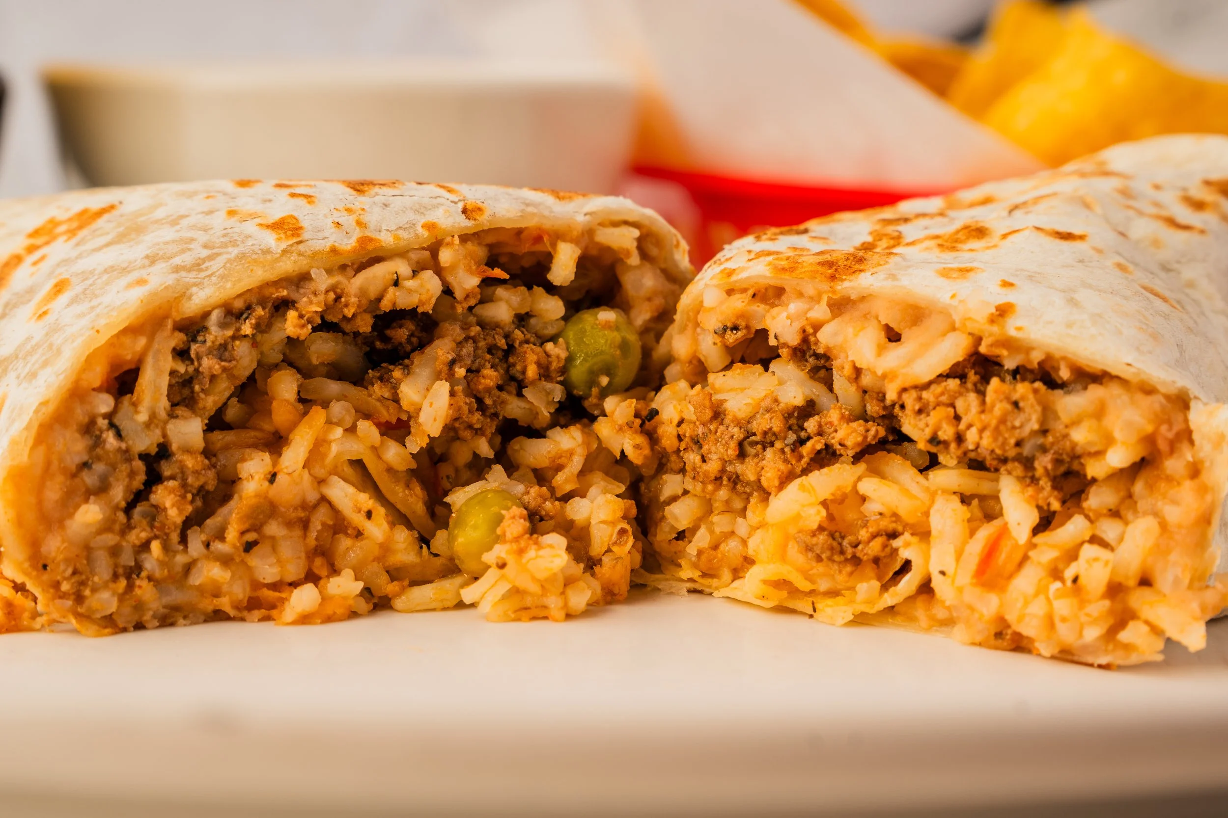 Cut in half burrito filled with ground beef, rice, and vegetables on a white plate with side dishes in background.