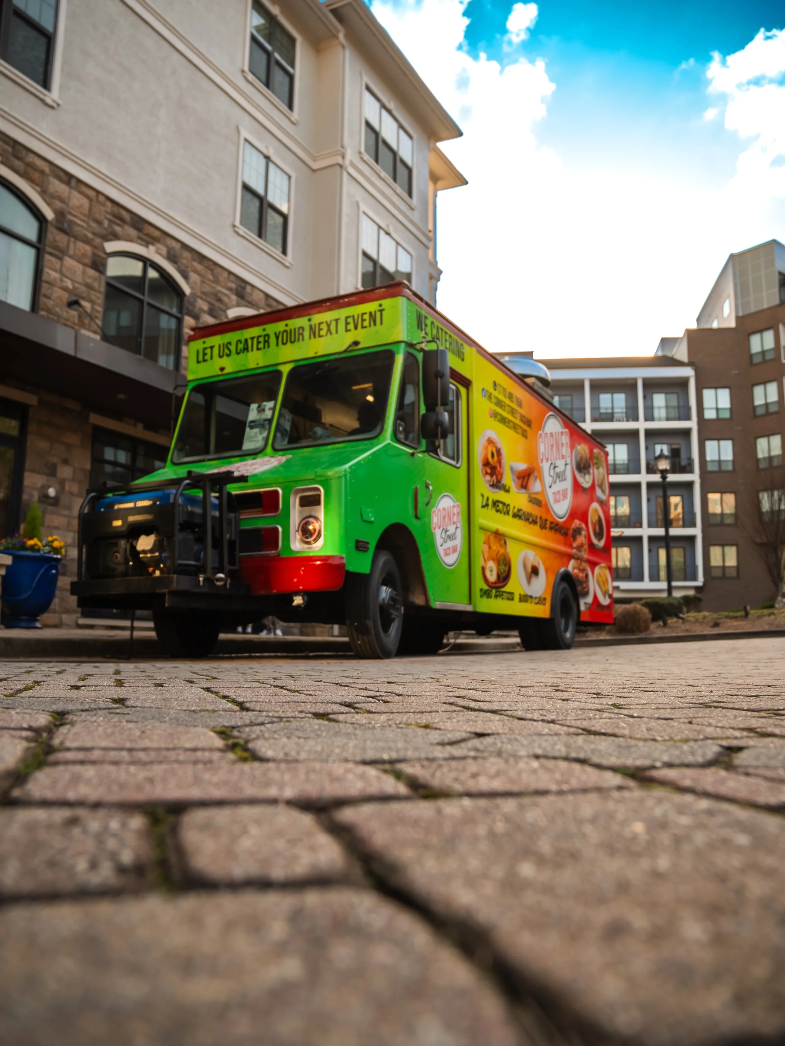 A colorful food truck parked on a brick sidewalk in front of modern multi-story buildings under a partly cloudy sky.