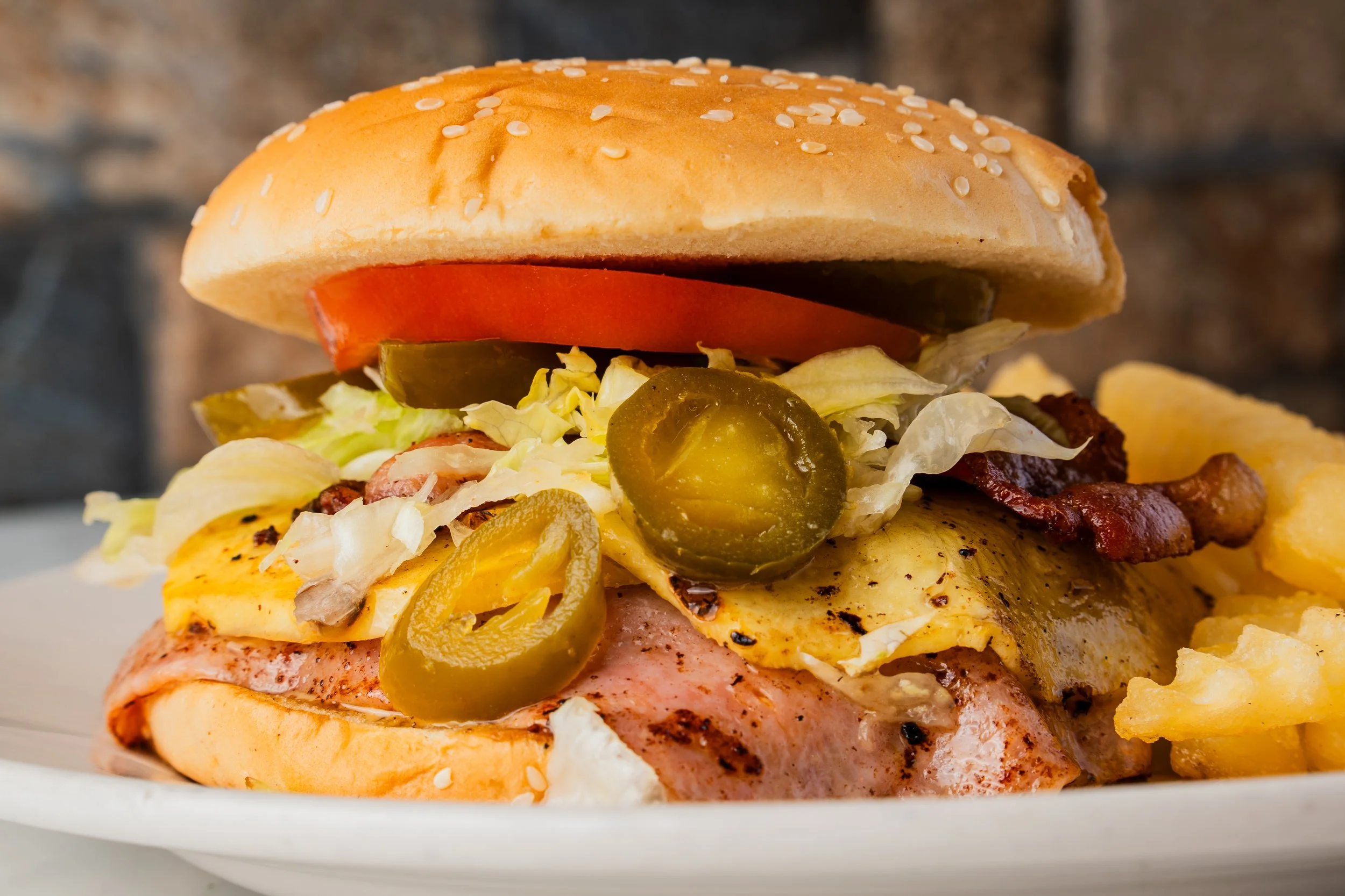 Close-up of a loaded burger with lettuce, tomato, pickles, fried egg, bacon, and potato chips on a plate, with a brick wall background.