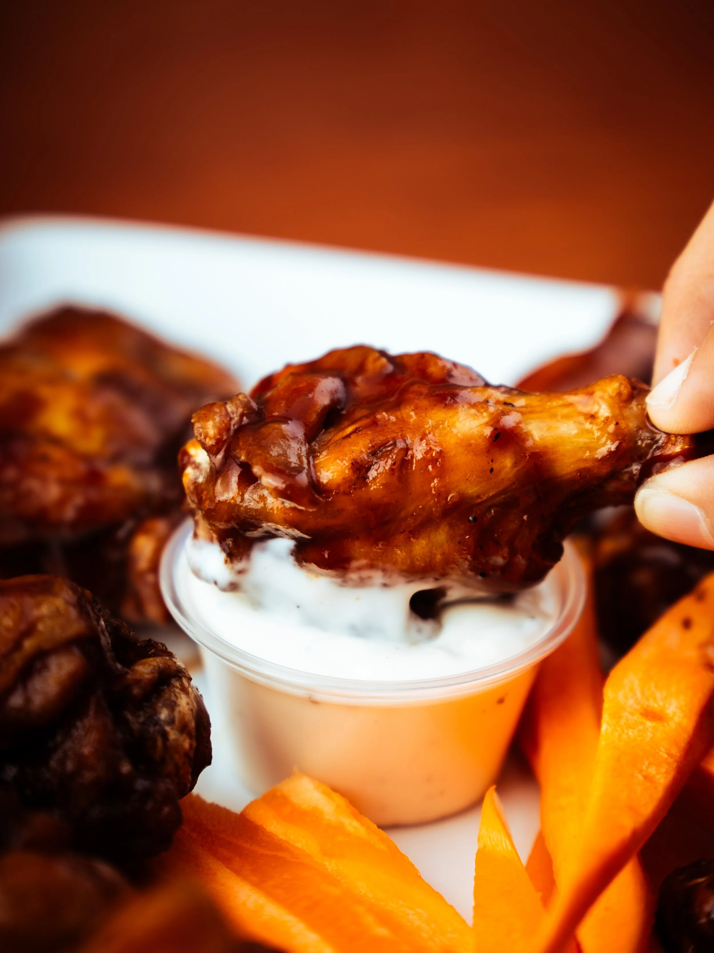 Close-up of a person's hand dipping a glazed chicken wing into a cup of ranch or blue cheese dressing, with additional chicken wings and carrots around the cup.