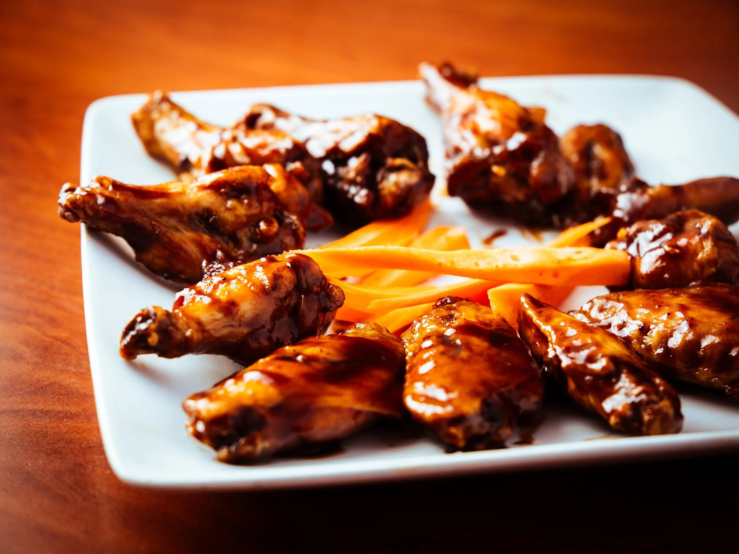 A white square plate with barbecue chicken wings and strips of carrots on a wooden surface.