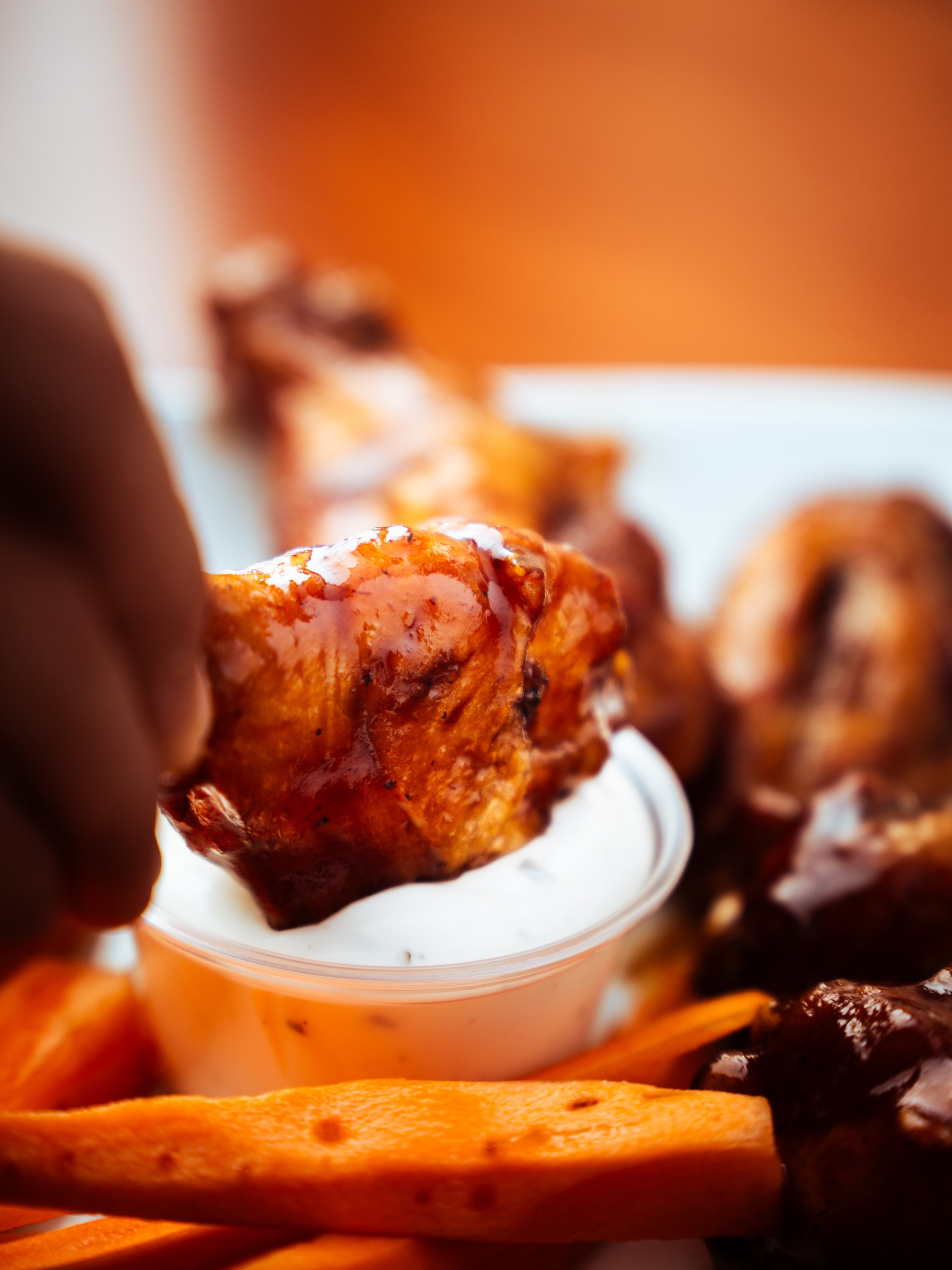 Close-up of a hand dipping chicken wing into a small container of ranch dressing, with other chicken wings and carrots on the plate in the background.