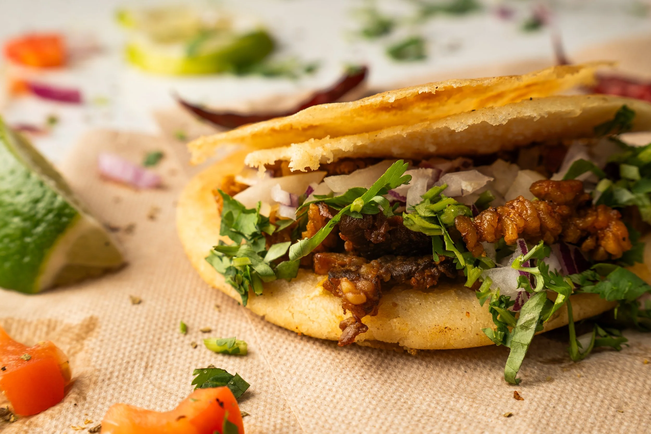 Close-up of a Gordita with lettuce, chopped onions, and cilantro, served with lime wedges and diced tomatoes.