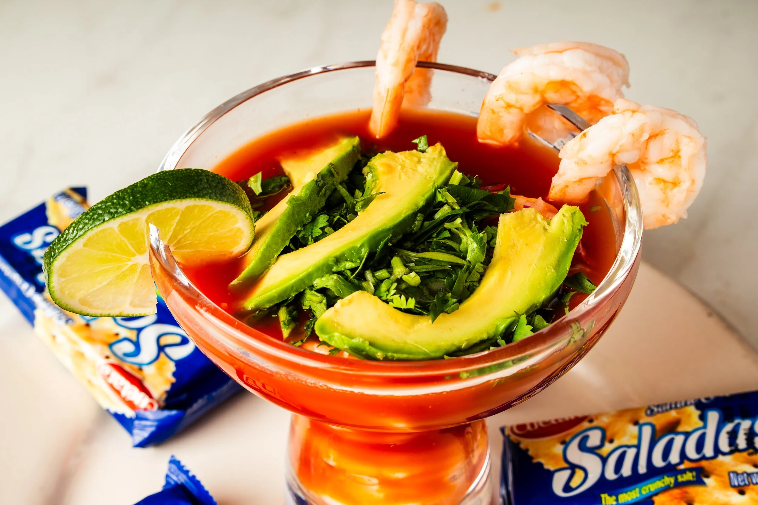 Shrimp cocktail in a glass bowl garnished with lime wedge, avocado slices, diced green herbs, and shrimp on skewers, with packets of saltine crackers around it.