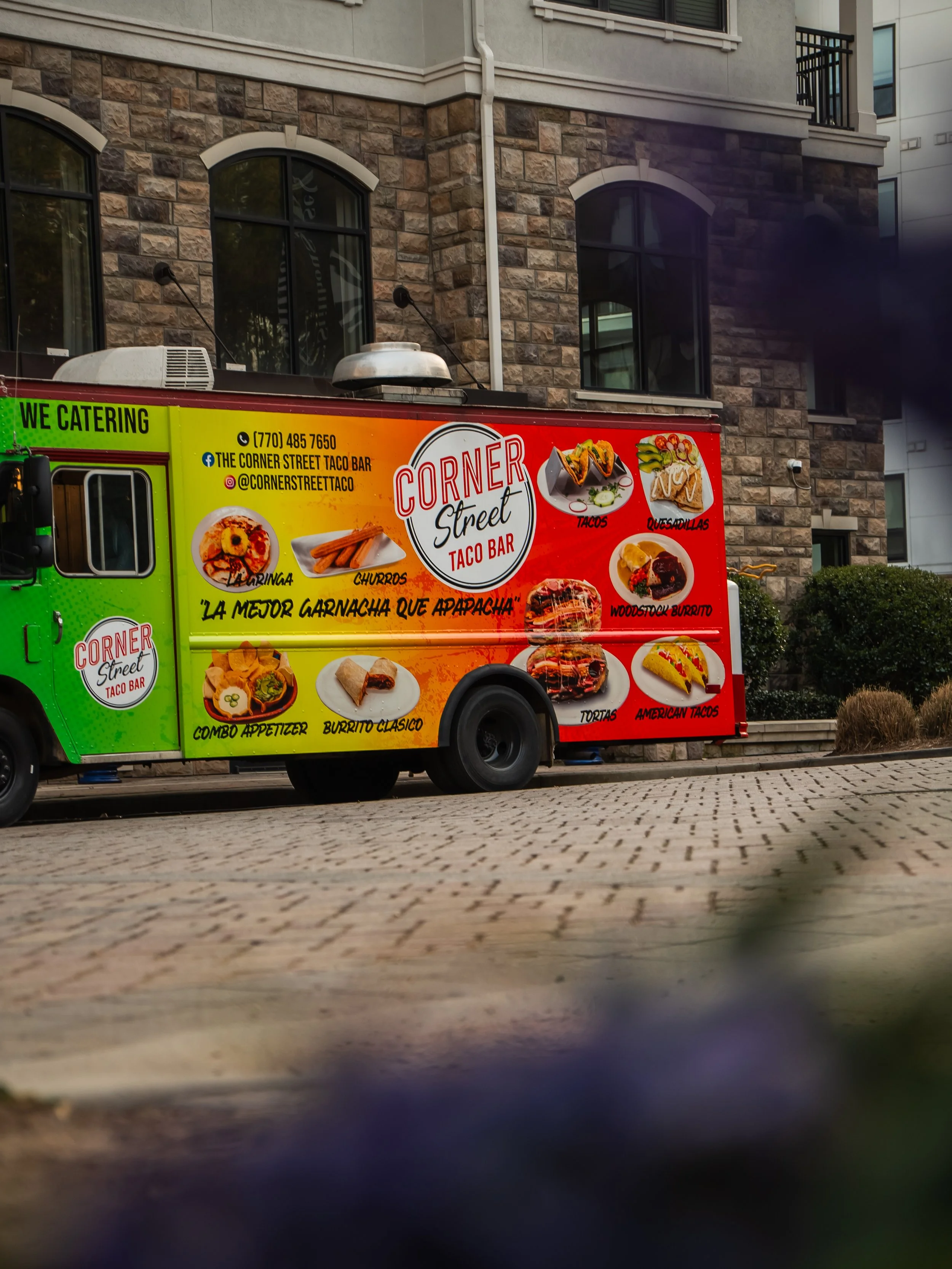 Colorful street taco truck parked on a brick sidewalk in front of a modern building with stone and white walls, advertising various Mexican foods and drinks.