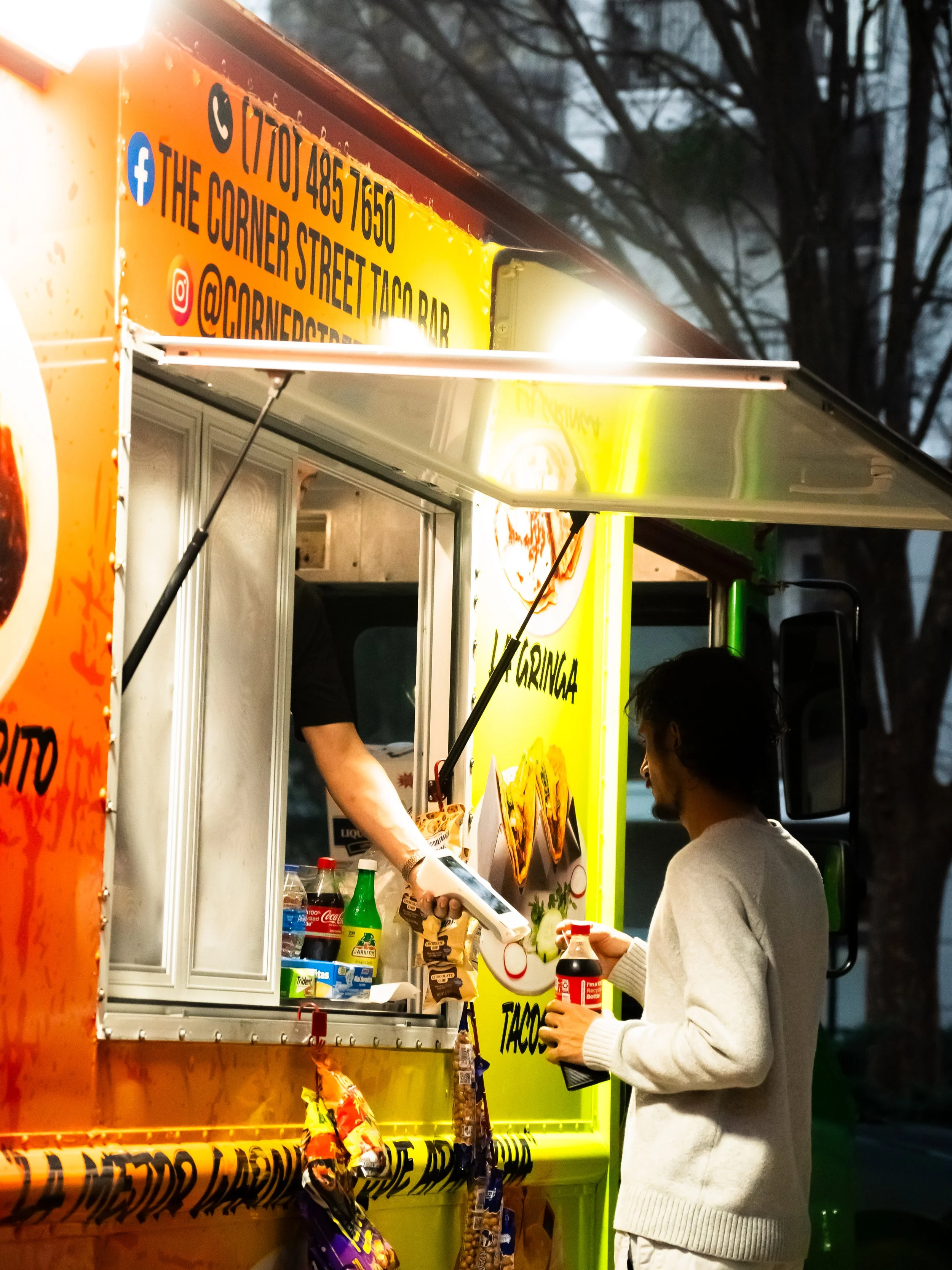 A person buying tacos from a brightly colored food truck with a customer service window, various bottles, and snack bags hanging below.