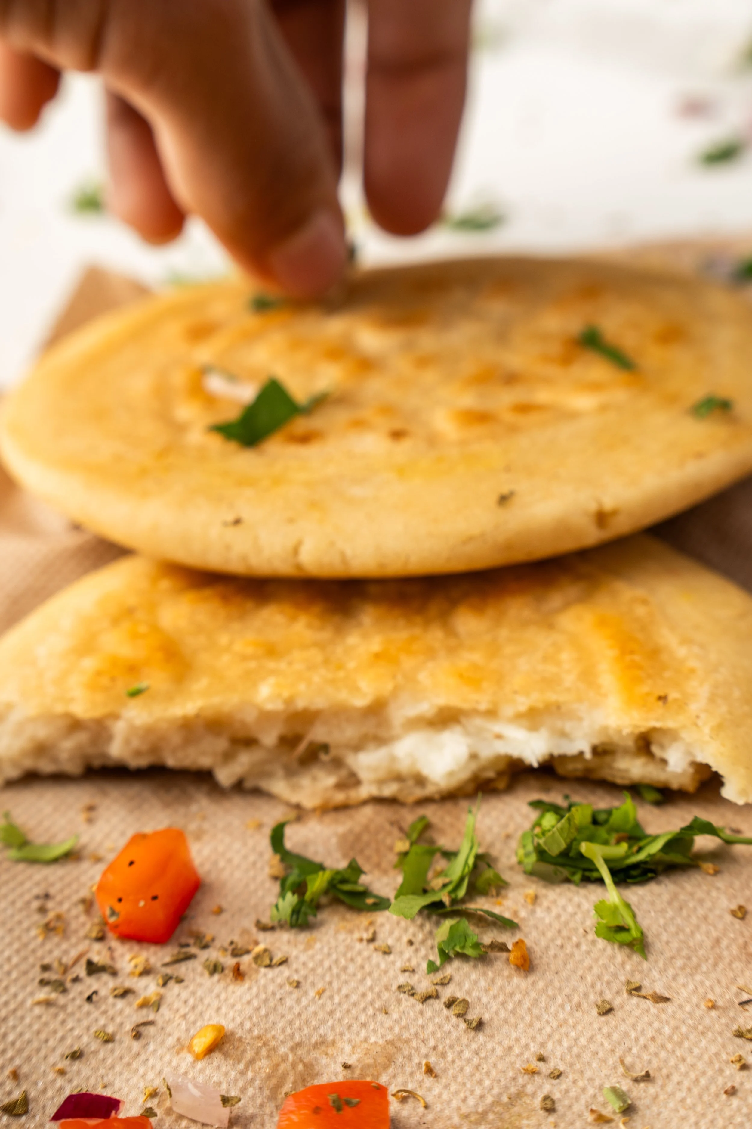Close-up of a person adding herbs to a pupusa with visible toppings, on a piece of parchment paper with chopped vegetables and herbs scattered around.