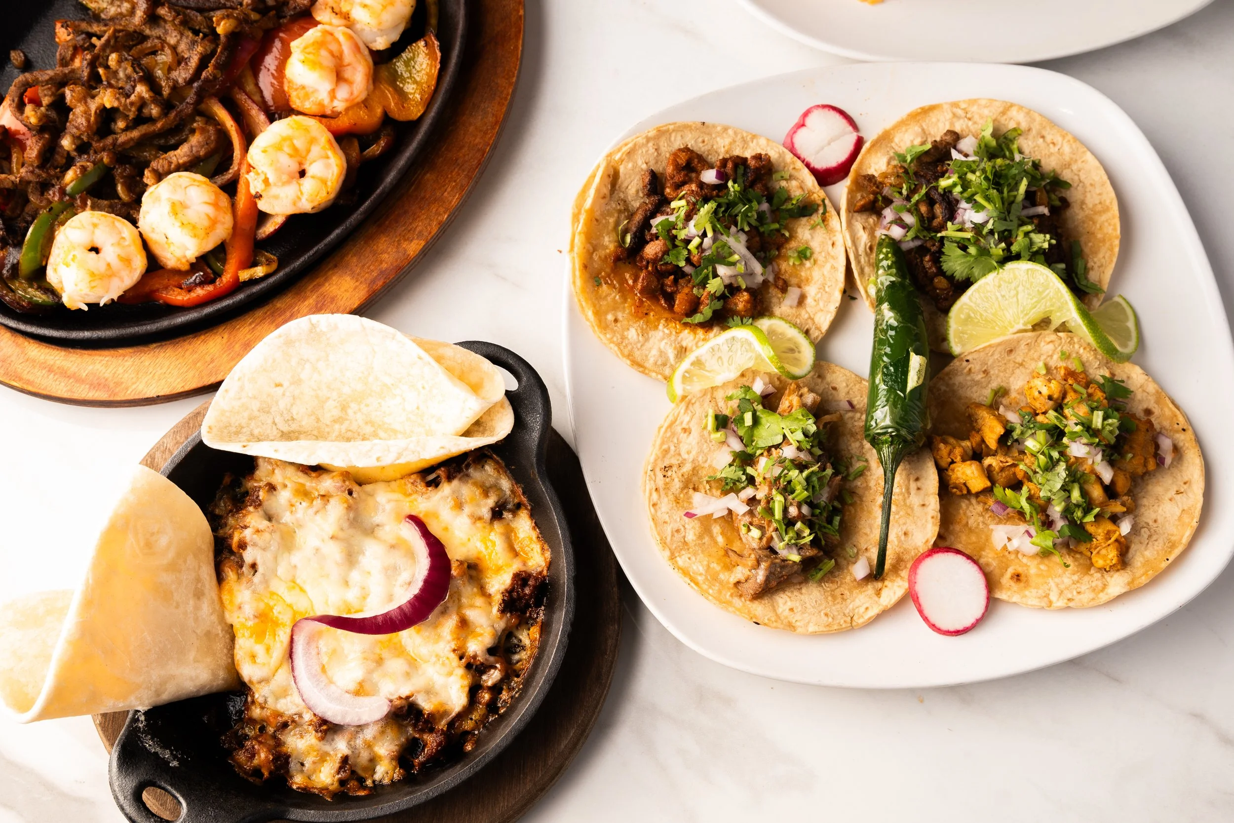 Plate of four tacos with meat, herbs, lime wedges, radish slices, served with a spicy chili pepper, a cast iron dish of melted cheese with onion slices and tortilla chips, and a wooden plate of stir-fried noodles with shrimp and vegetables.
