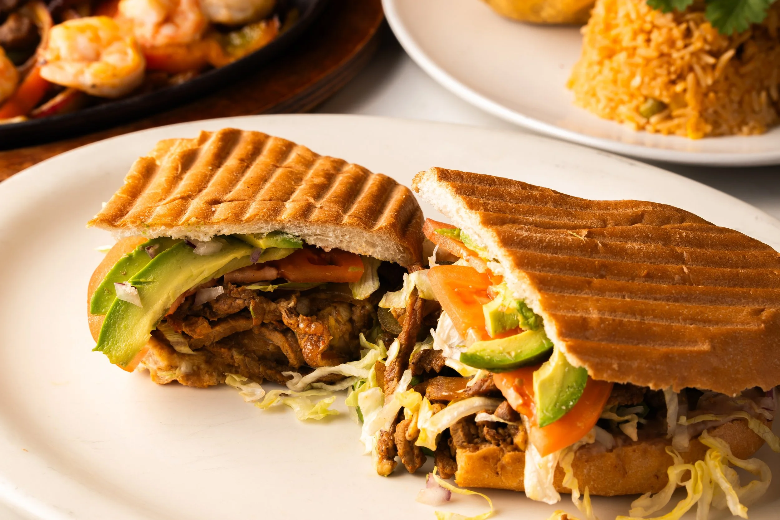 Torta cut in half on a white plate, filled with avocado, shredded lettuce, sliced tomato, grilled meat, and vegetables, with toasted bread grill marks.