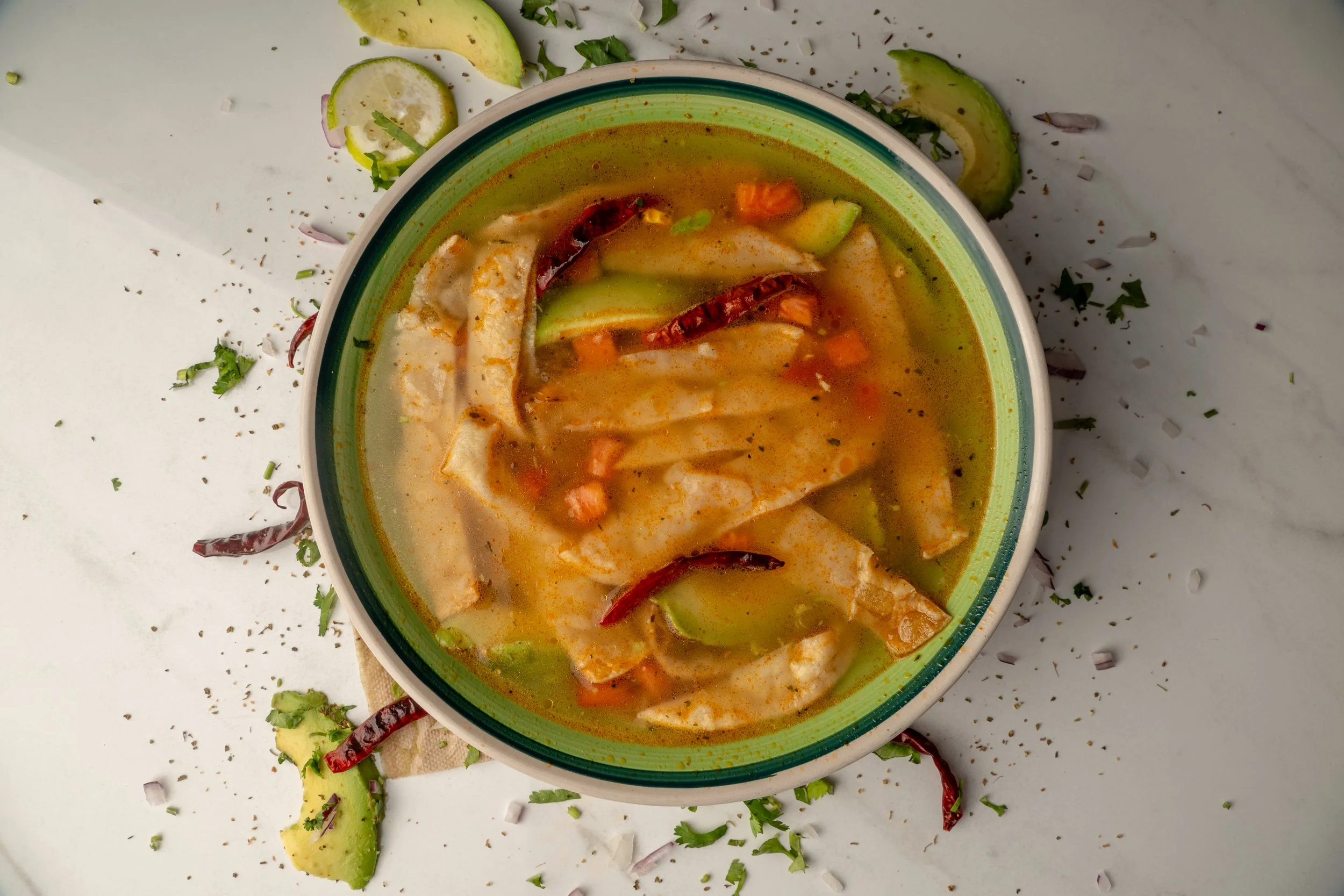 A bowl of Caldo de Azteca with vegetables, chili peppers, and herbs, with lime slices, avocado, and chopped herbs scattered around on a white surface.