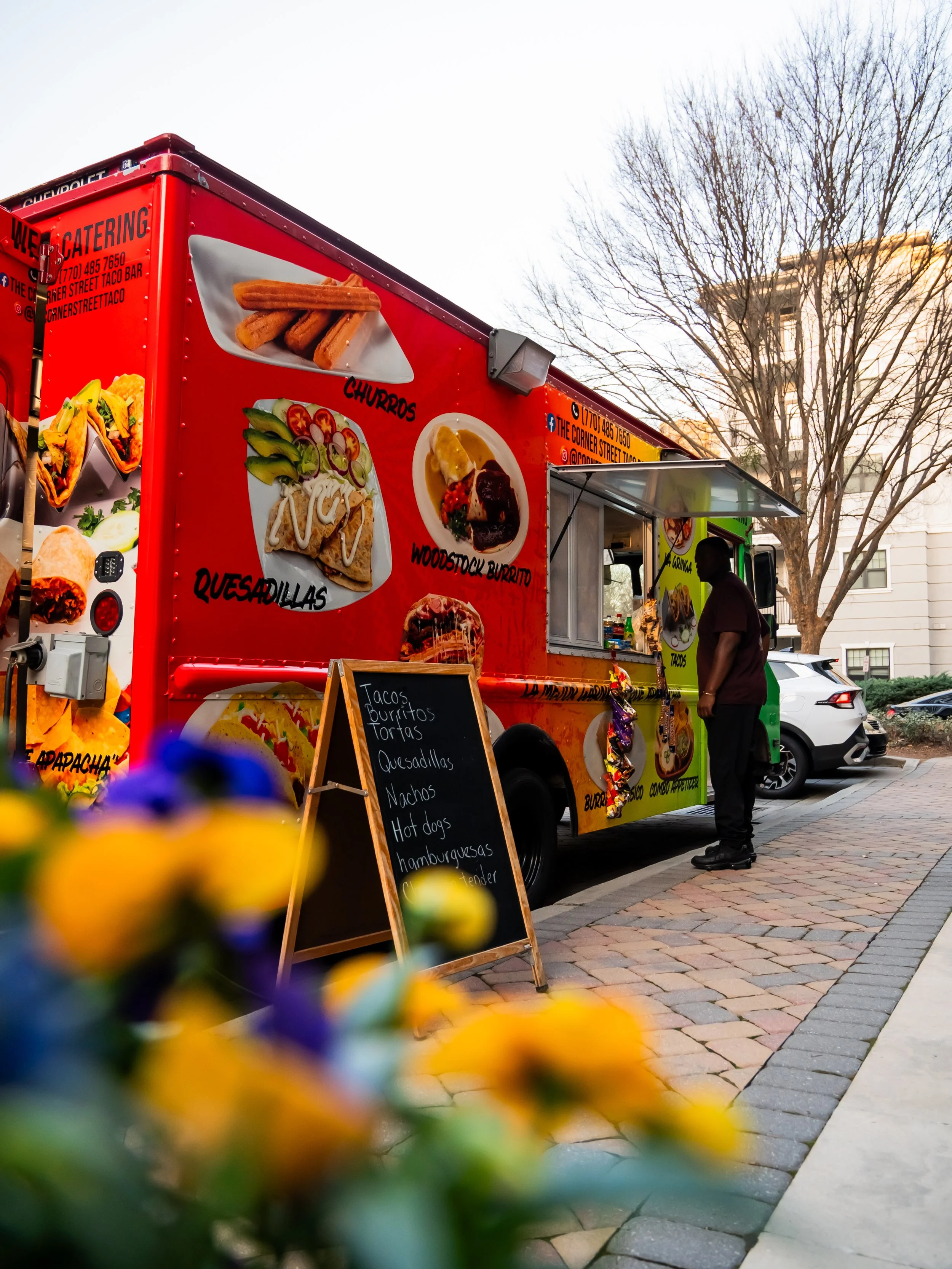 A colorful food truck parked on a brick sidewalk with a man standing nearby. The truck displays images of churros, quesadillas, and a buffalo burger. A chalkboard sign in front lists menu items including tacos, burritos, tortas, quesadillas, nachos, 