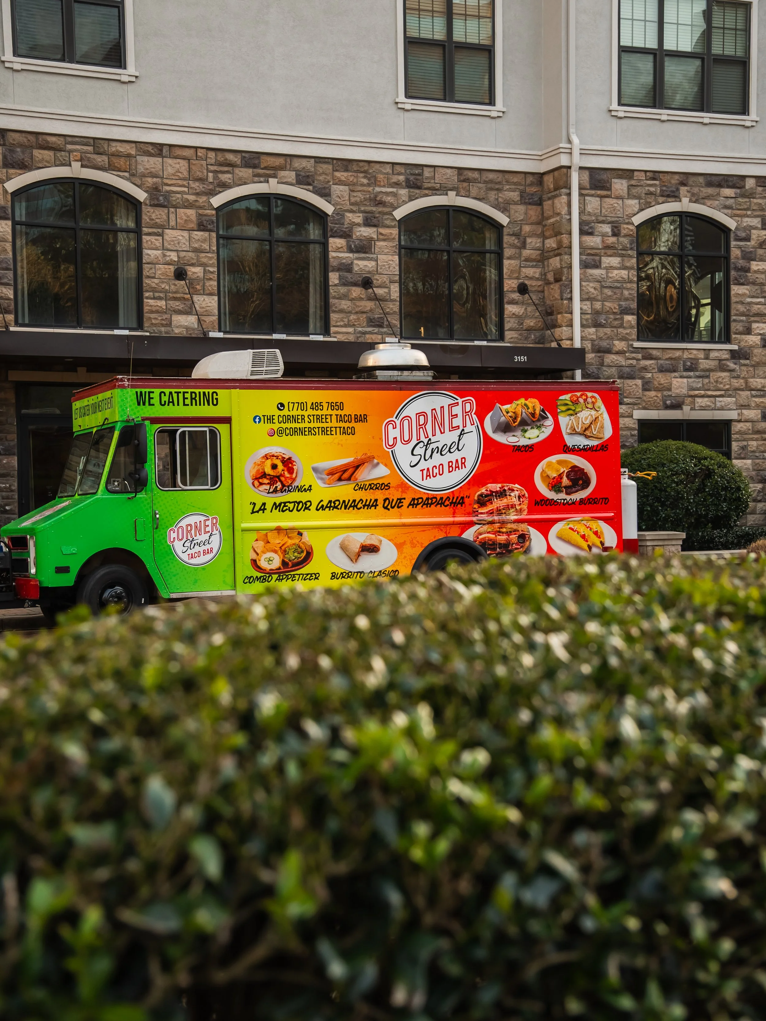 Green and red Taco Truck parked in front of a building with stone and stucco exterior, advertising Mexican food including tacos, quesadillas, burritos, and more.