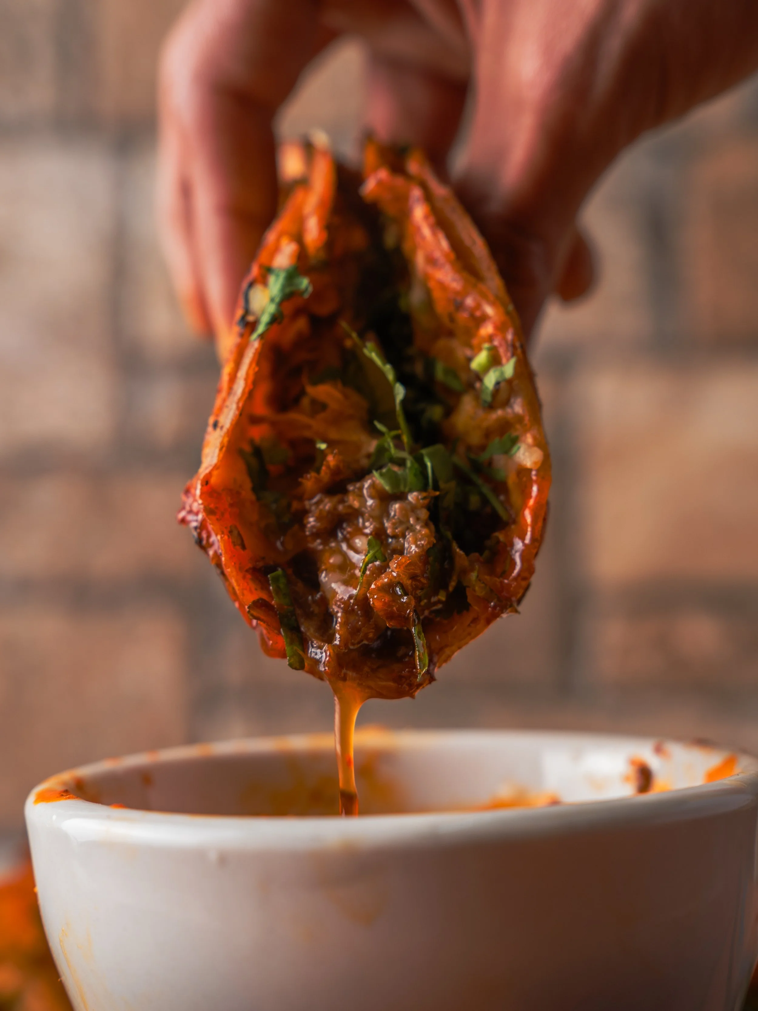 A hand holding a Birria taco being dipped into a white bowl of consome.