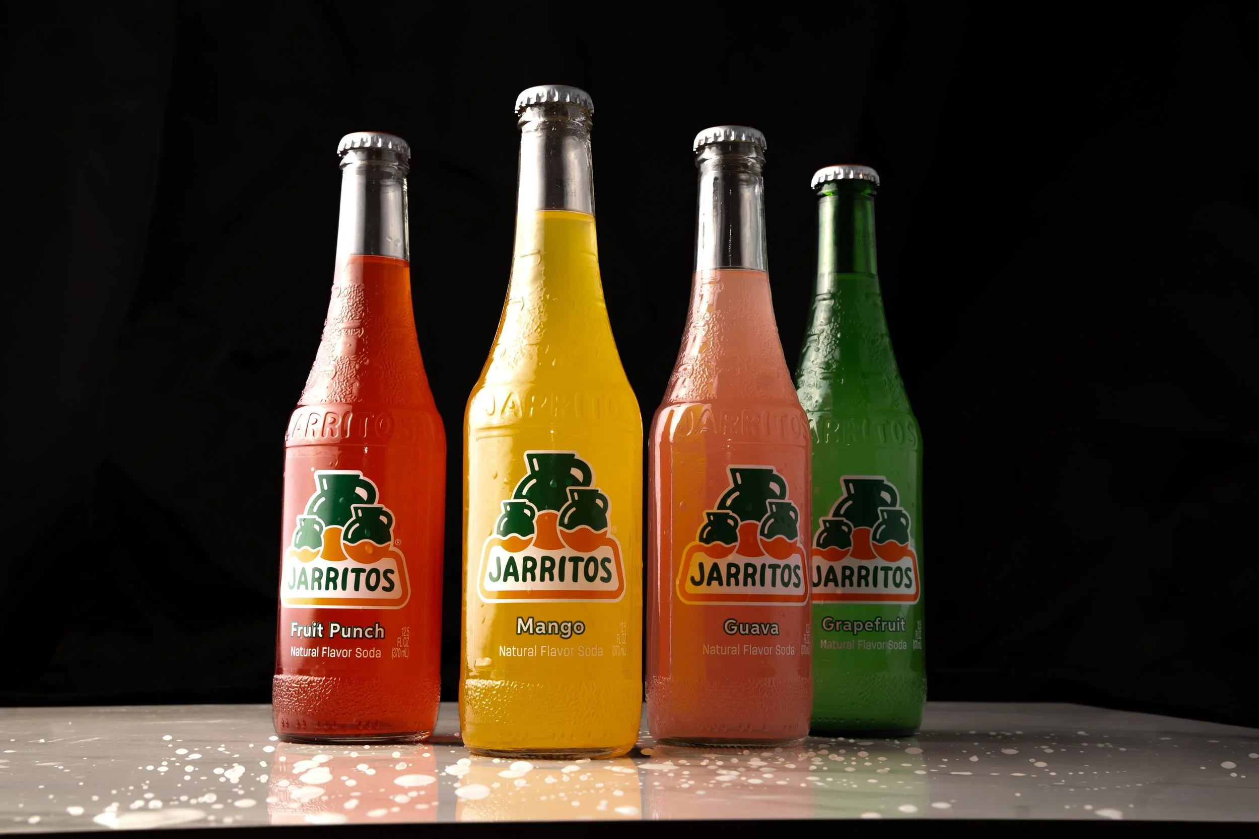 Four glass bottles of Jarritos soda flavors on a table with white splashes, against a black background. From left to right: Fruit Punch, Mango, Guava, and Grapefruit.