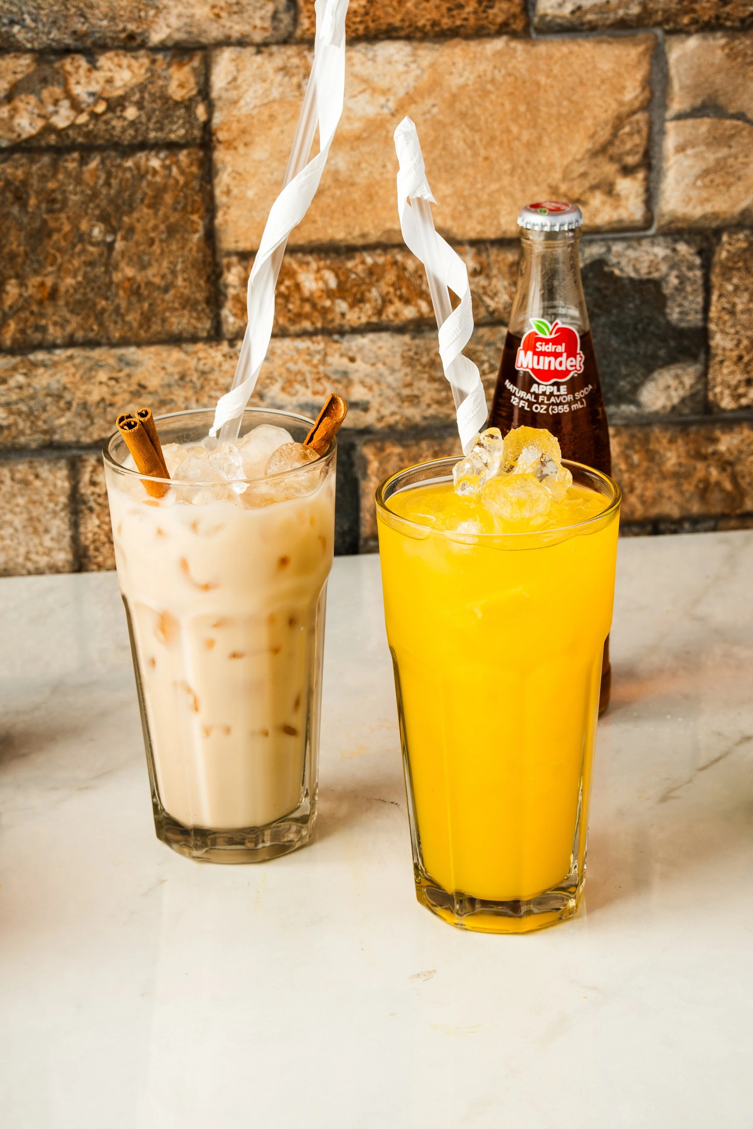 Two tall glasses of beverages, one with horchata topped with cinnamon sticks and a paper straw, and the other with an agua Fresca and ice, with a bottle of mexican coke in the background against a brick wall.