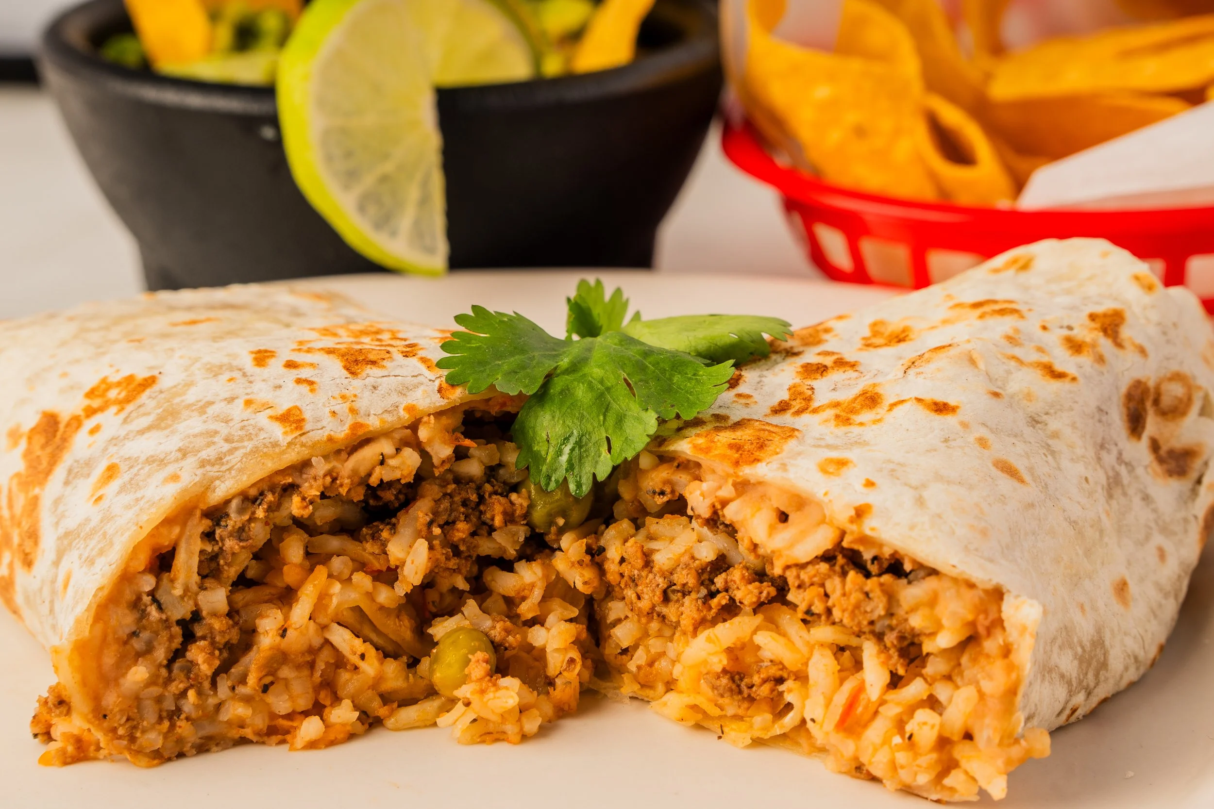 Cross-section of a burrito filled with rice, ground meat, beans, and cheese, garnished with cilantro, with lime slices, chips, and salsa in the background.
