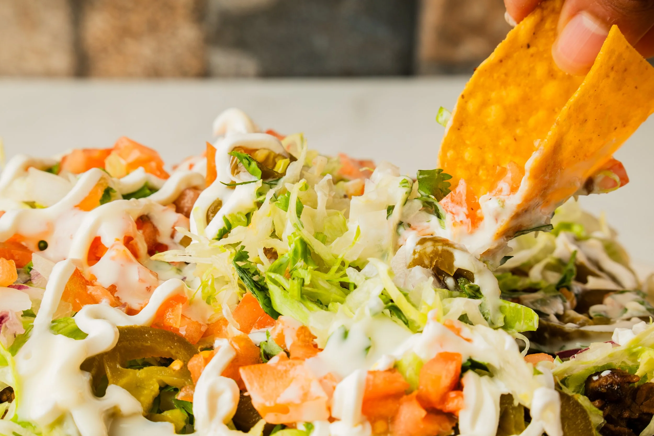Toppings being added to a taco salad, including shredded lettuce, chopped tomatoes, sliced jalapeños, shredded cheese, and sour cream.