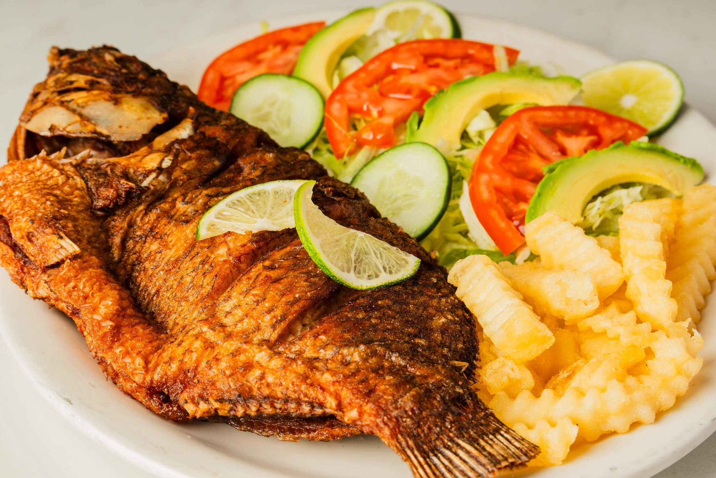 Plate with cooked whole fish garnished with lime slices, sides of crinkle-cut fries, and a salad of sliced cucumbers, tomatoes, lettuce, avocado, and lemon.