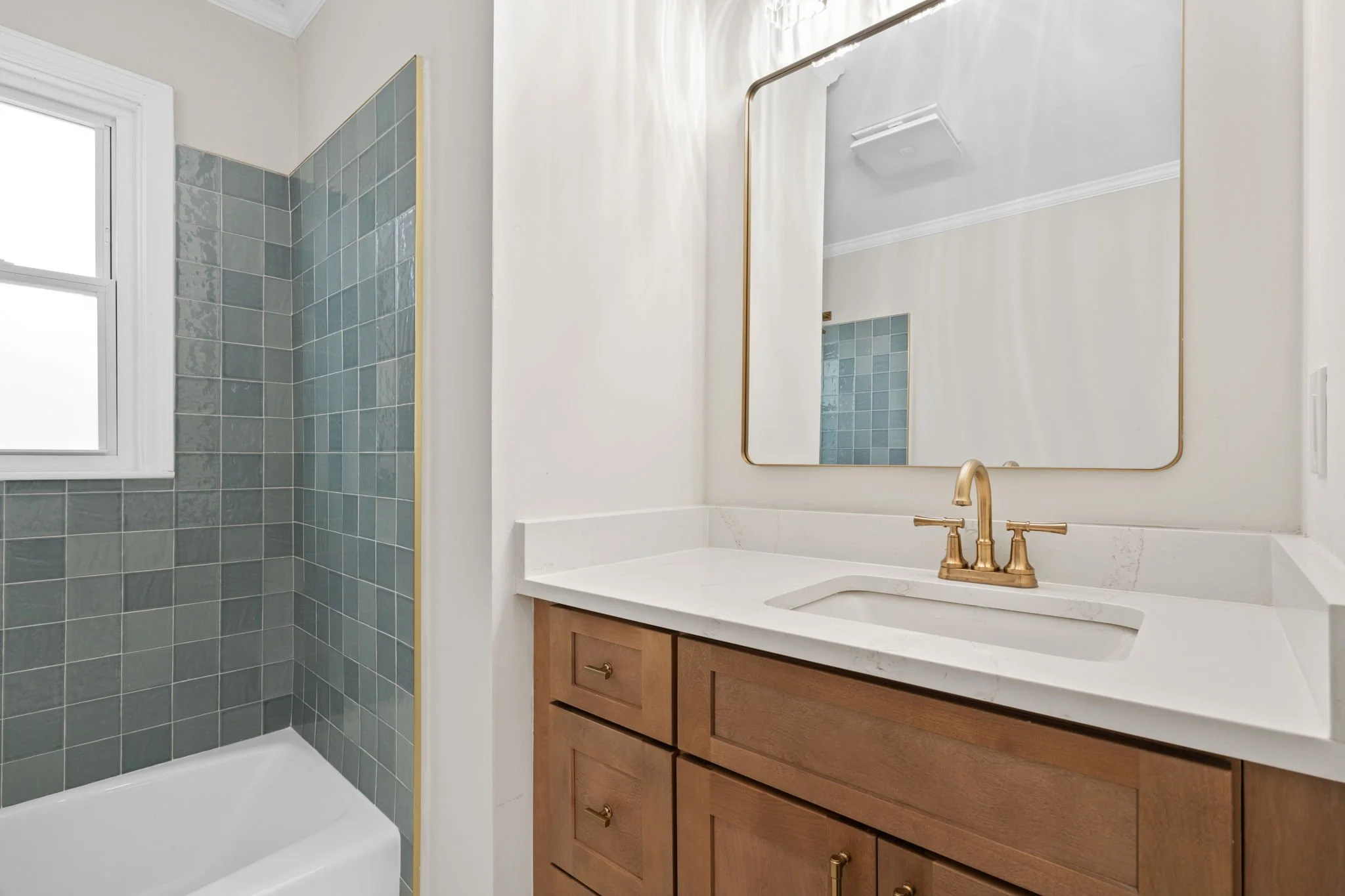 Bathroom with a wooden vanity, white countertop, brass faucet, large mirror, and walk-in shower with blue tiles and a window.
