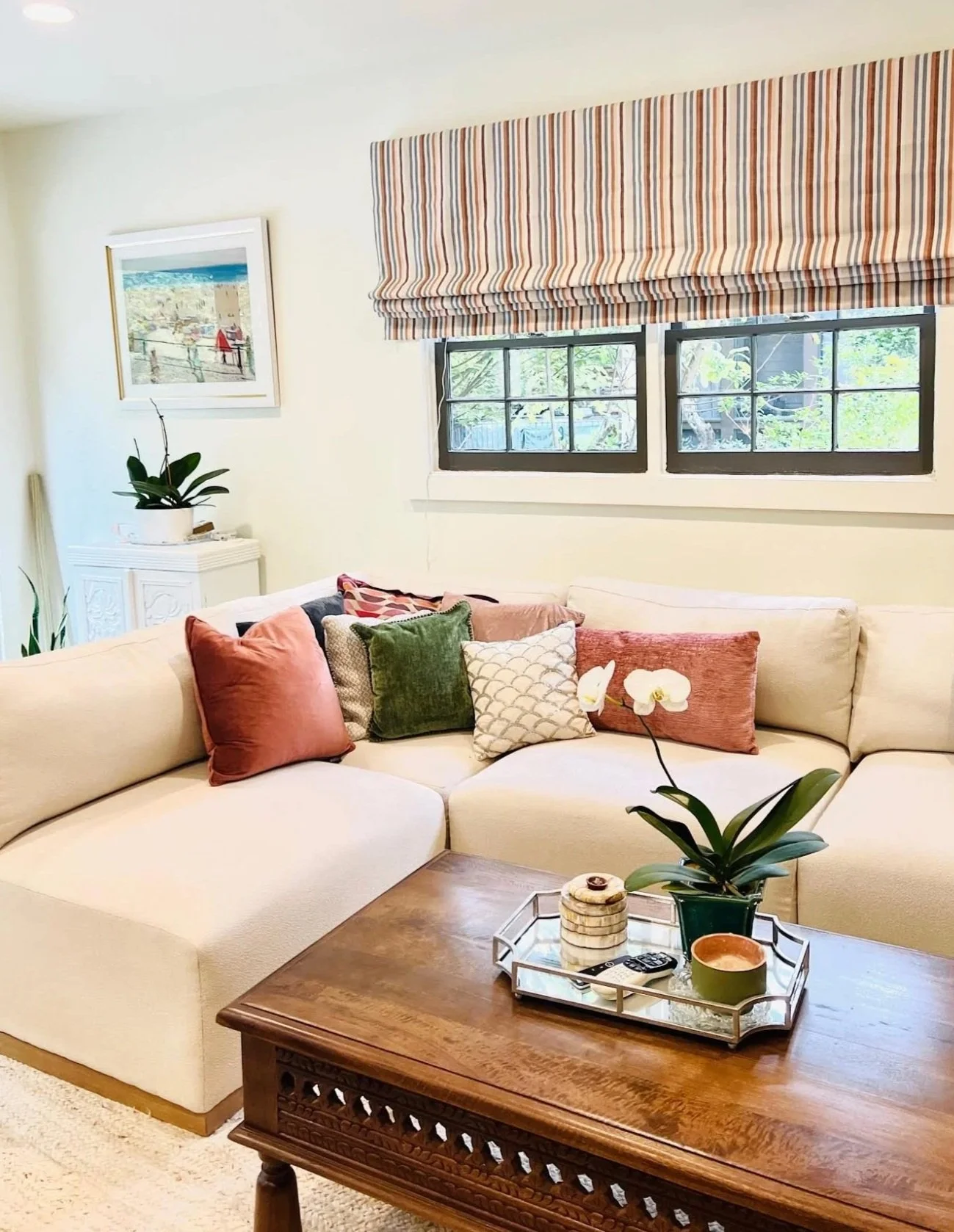 Living room with a white sectional sofa decorated with colorful cushions, a wooden coffee table with decorative items and a potted orchid, a window with striped valance, a piece of wall art, and houseplants.