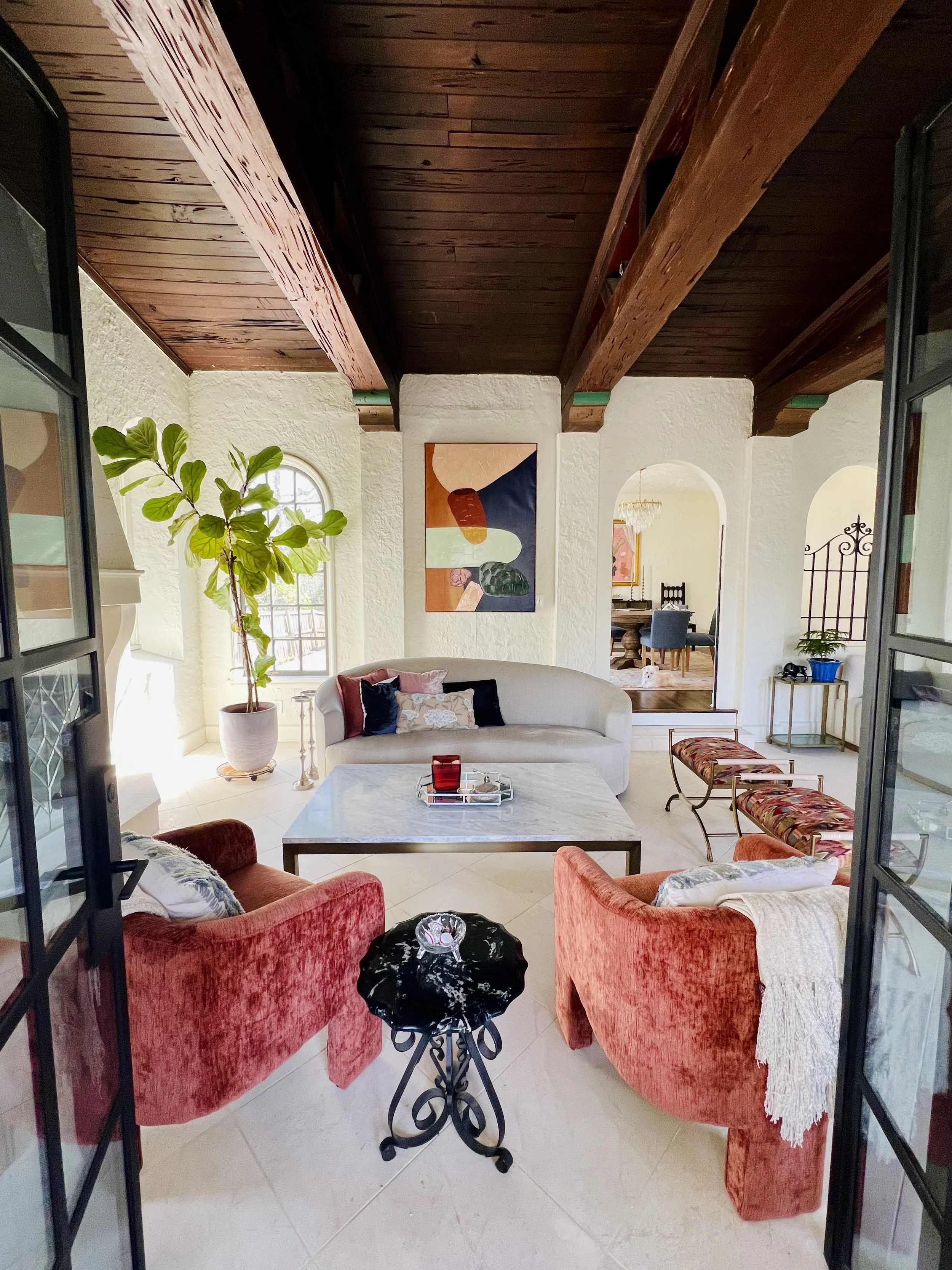 Living room with two pink velvet armchairs, a black side table, a large white curved couch with pillows, a marble coffee table, a large potted plant, and an abstract wall art. Open doorway leads to dining area with a chandelier and wooden table.