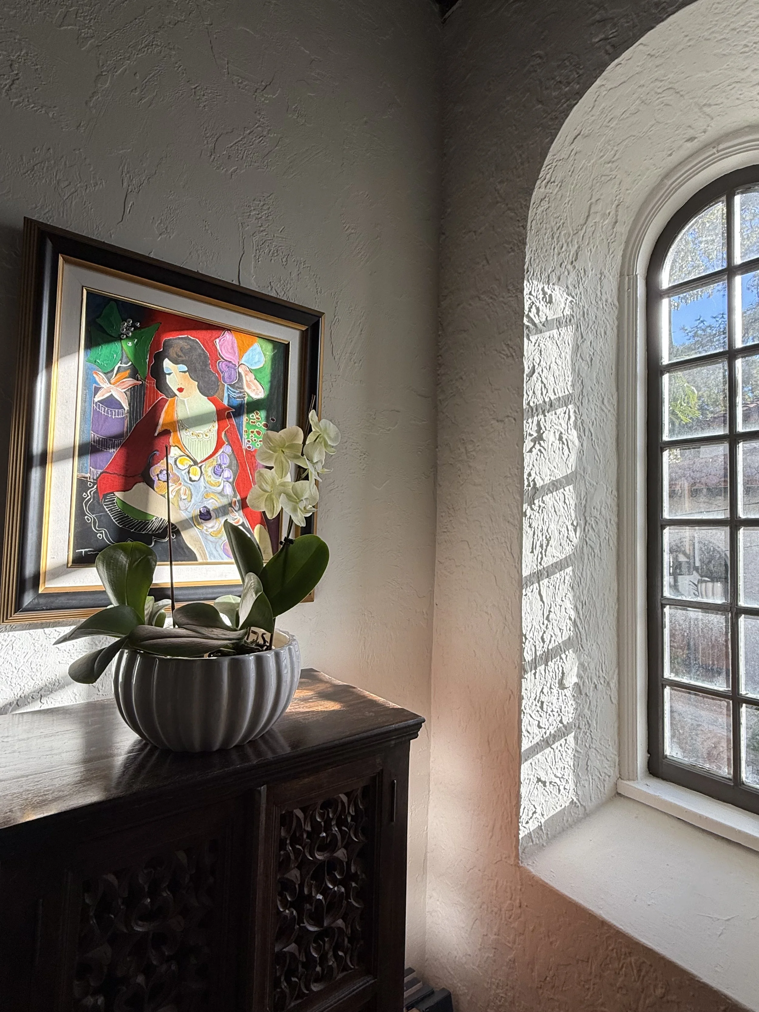 Sunlight streaming through a arched window illuminating a painting by Tarkay on a dark wooden cabinet with a colorful framed artwork on the wall behind.