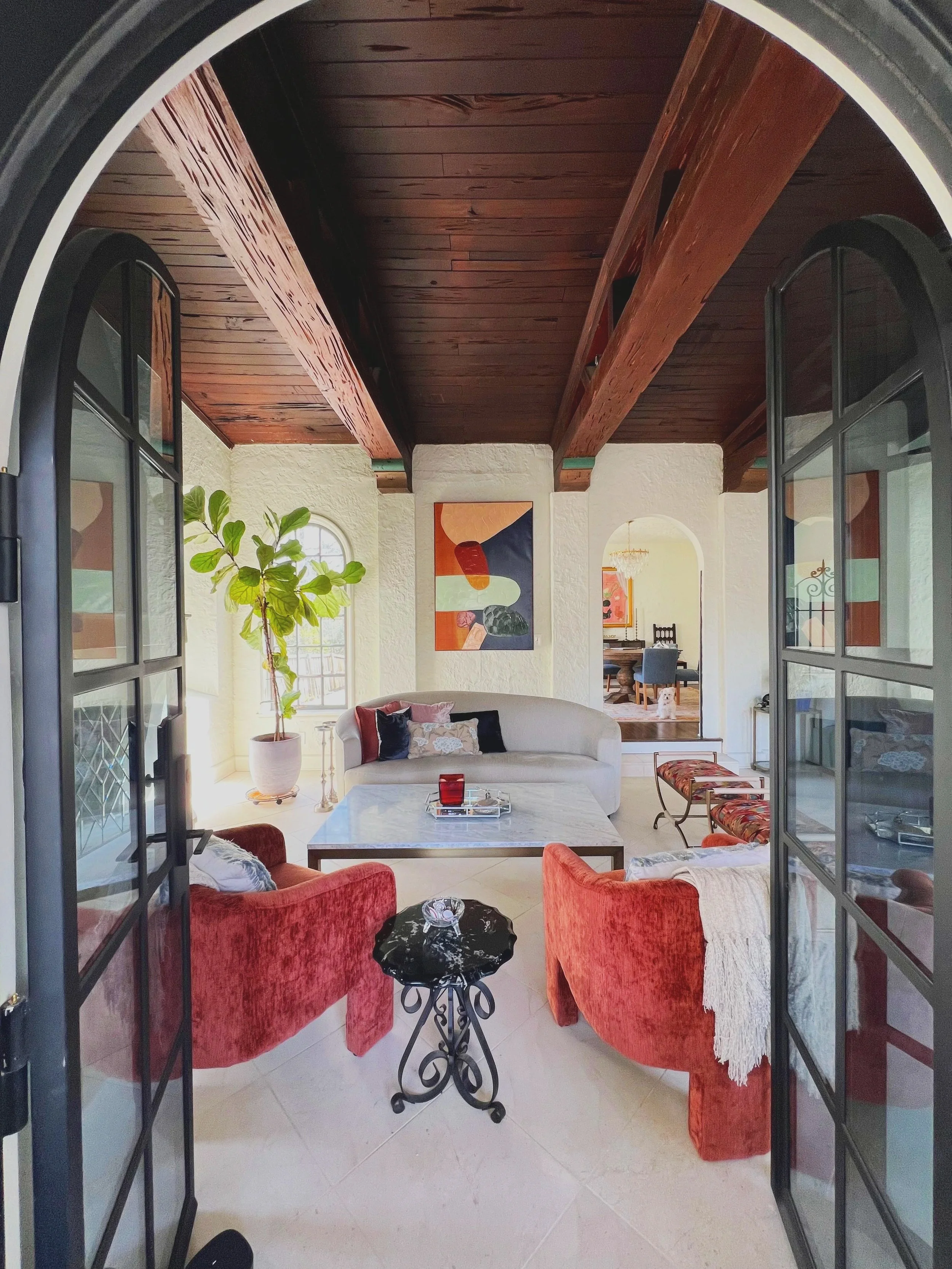 Living room with white sofa, red velvet chairs, a black side table, a coffee table with red and silver decor, a large potted plant, colorful abstract art, and an arched doorway leading to a dining area.